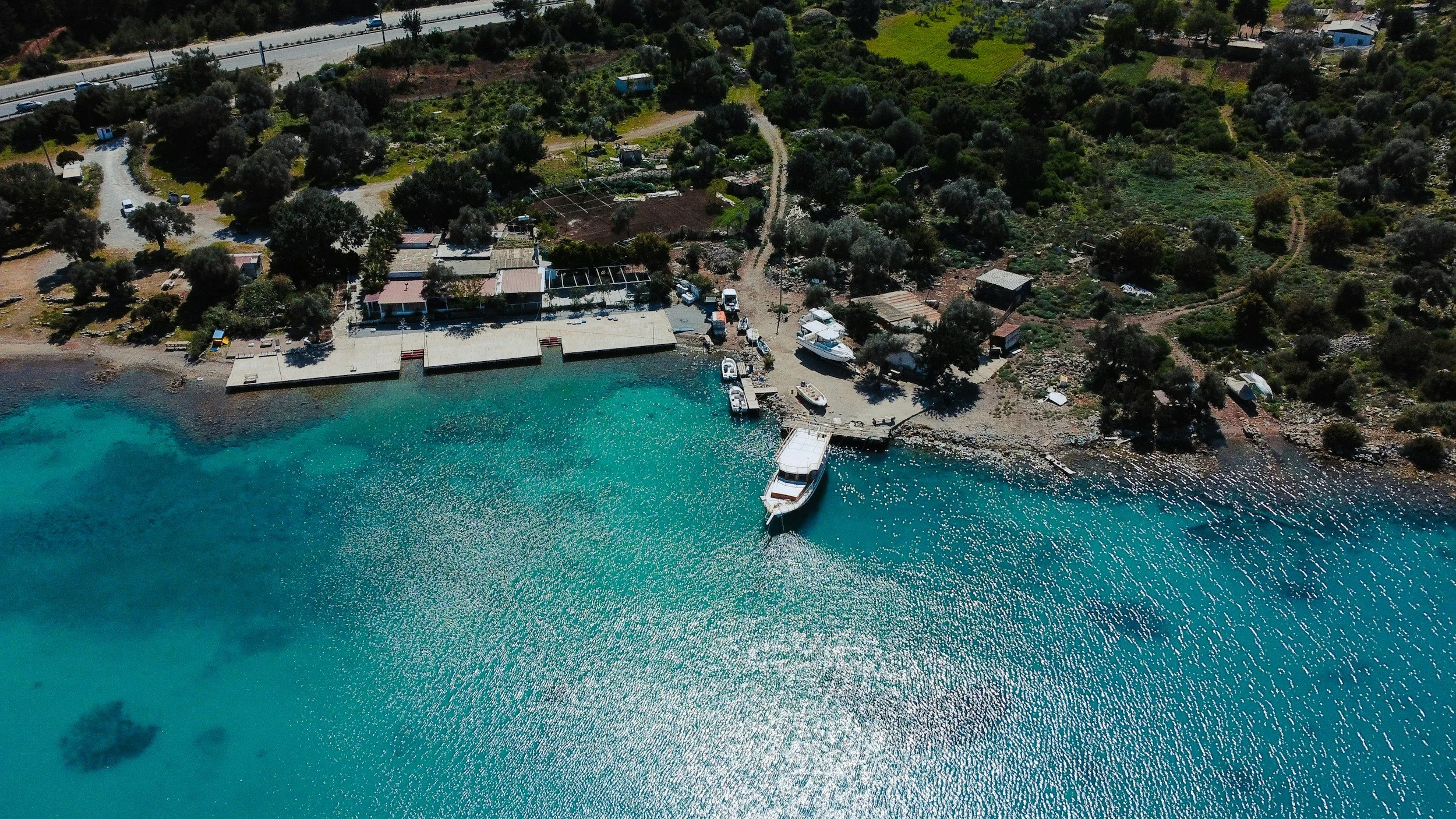 Aerial view of a small coastal area with turquoise water, boats docked at a pier, a grassy shoreline with trees, and a nearby road with vehicles.