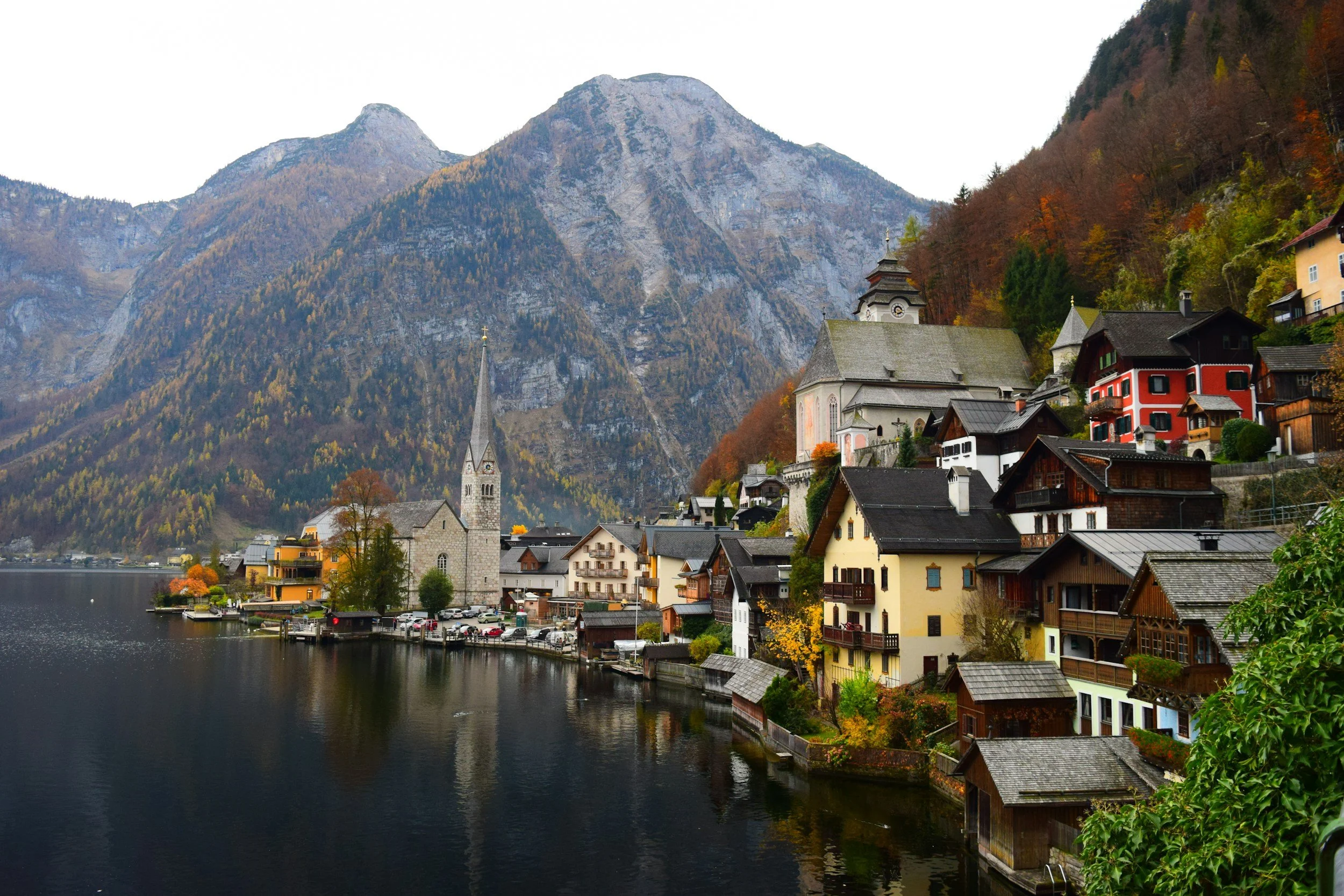 A picturesque lakeside village with colorful houses, a church with a tall steeple, surrounded by mountains and autumn foliage.