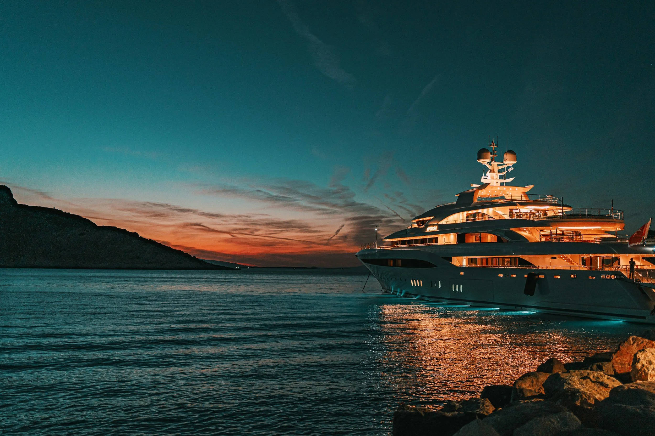 Luxury yacht docked at a rocky shoreline during sunset with colorful sky and hills in the background.