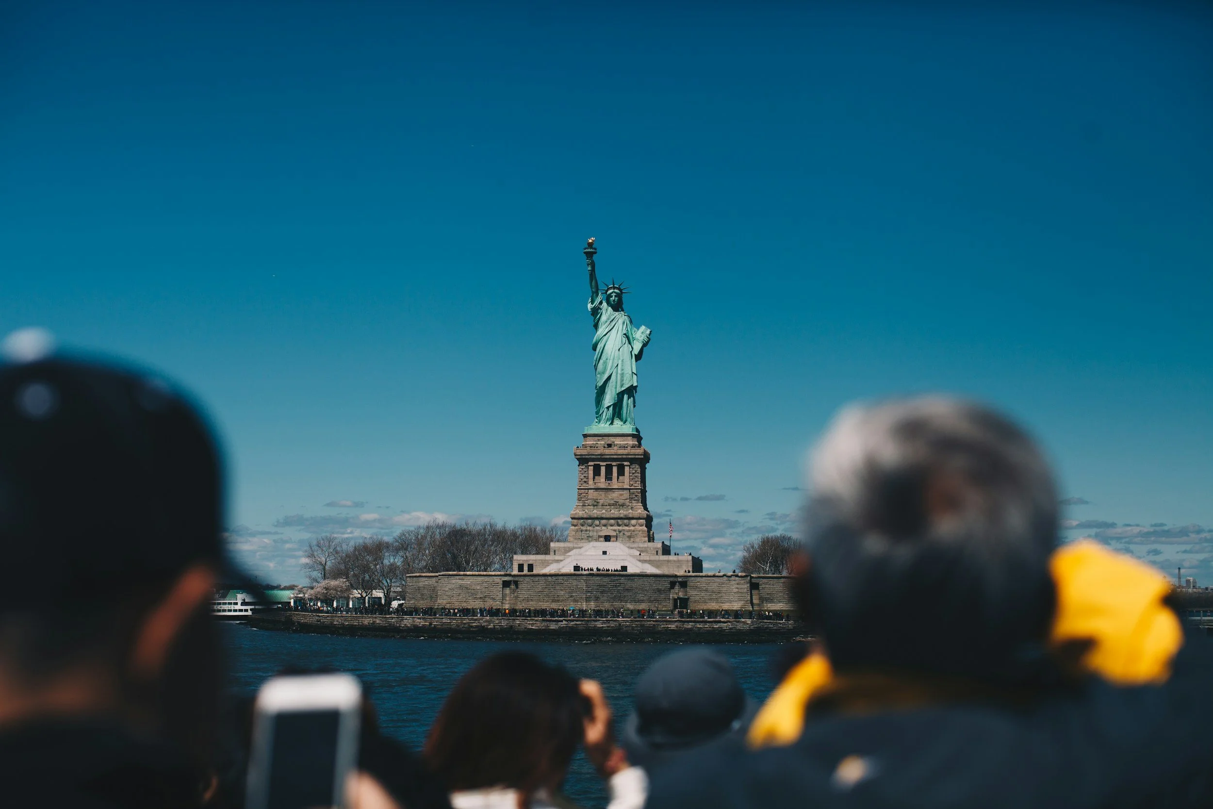 Tourists observing the Statue of Liberty on Liberty Island in New York Harbor with a clear blue sky in the background.