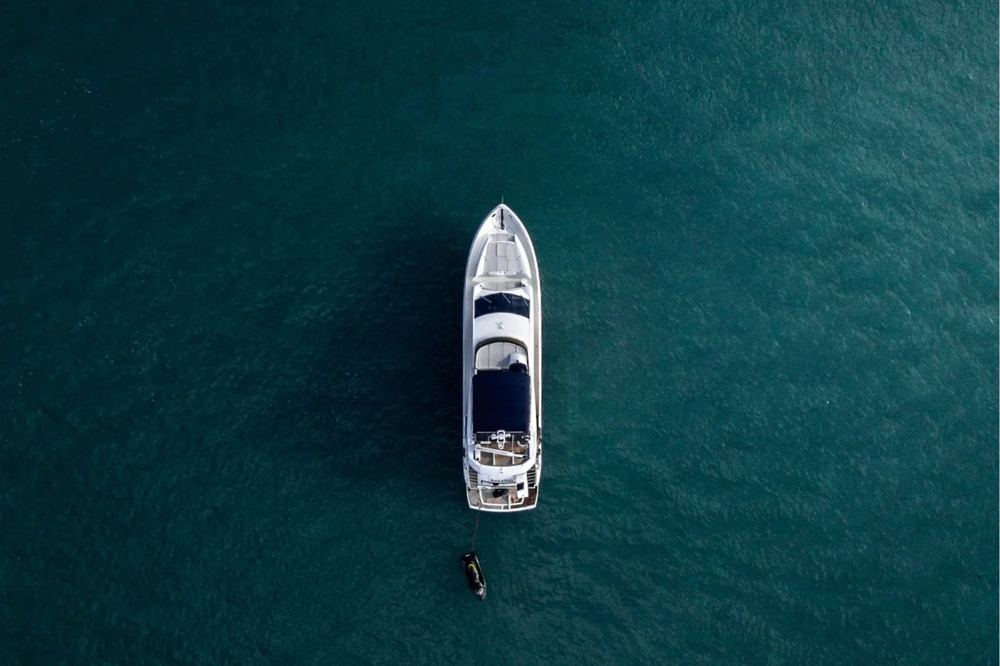Overhead view of a white yacht anchored in a calm, greenish-blue body of water, with a small black jet ski tethered to it.