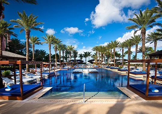 Luxury resort pool area with palm trees, lounge chairs, and cabanas on a sunny day.