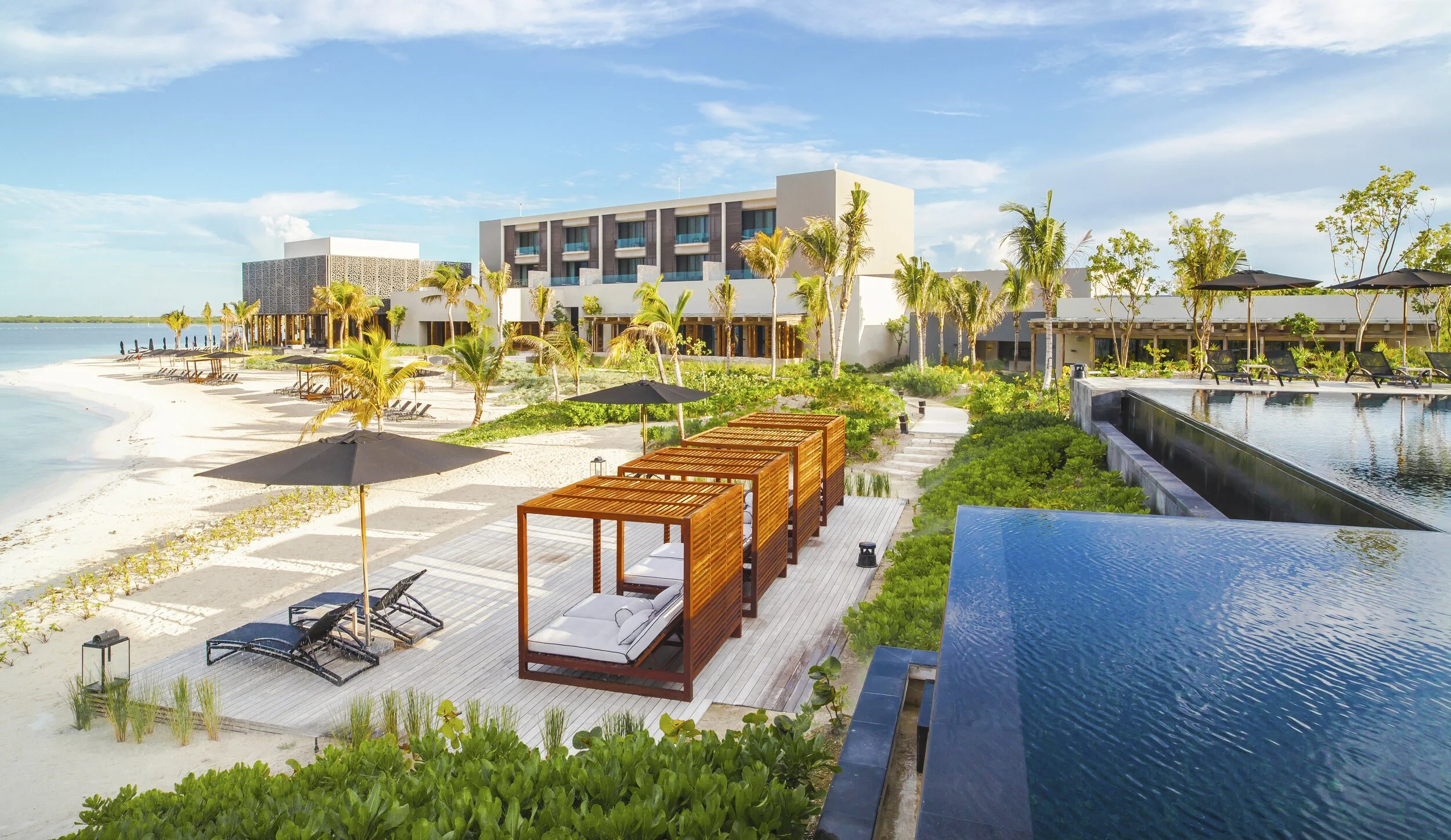 Luxury beachside resort with cabanas, lounge chairs, palm trees, and a swimming pool overlooking the ocean on a sunny day.