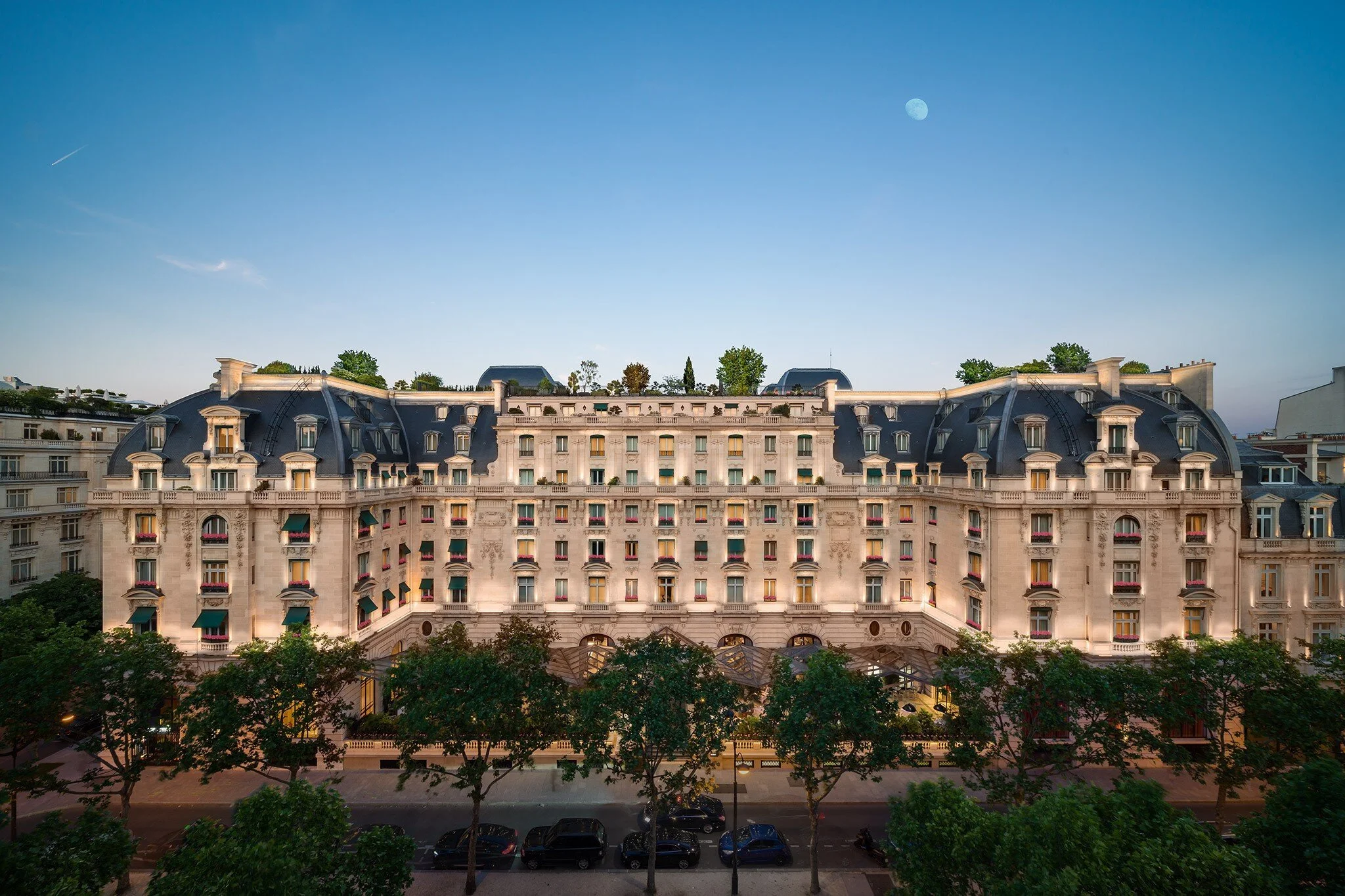 A grand, historic Parisian building with ornate architecture, multiple windows with a combination of awnings and balconies, and a mansard roof. The building is illuminated by evening light, with trees and parked cars along the street in the foreground, and a clear sky with the moon visible.