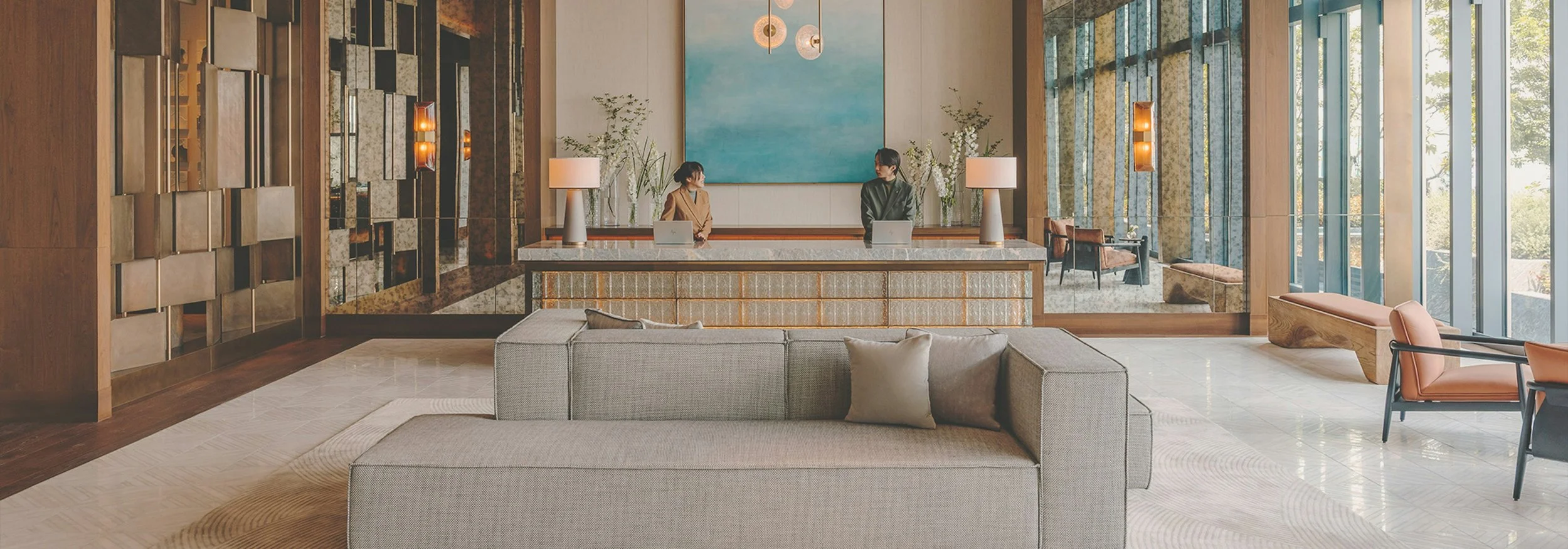 Hotel lobby with a reception desk, two staff members, seating area, large windows, and modern decor.