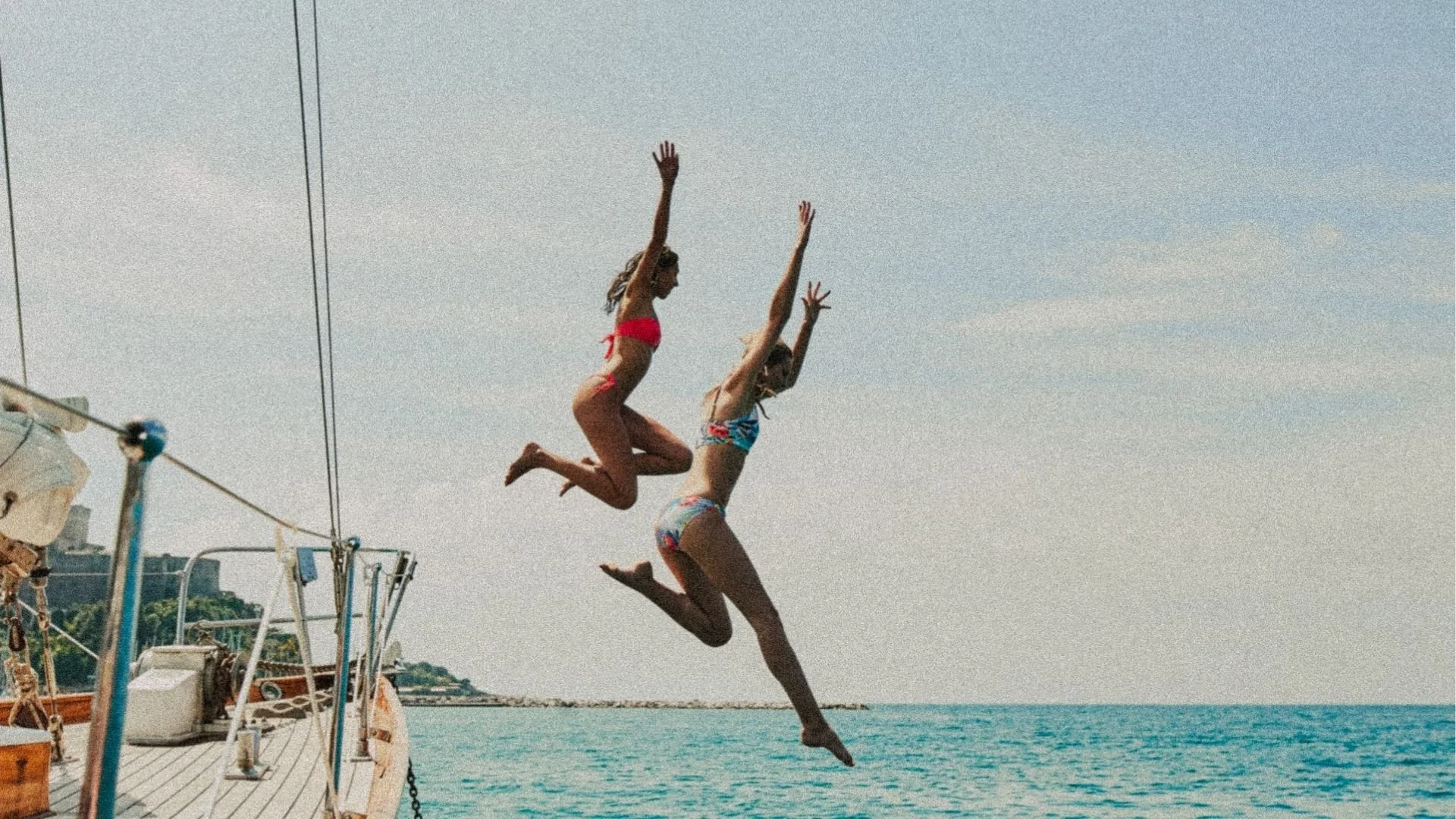 Two women in swimsuits jumping into the water near a boat on a sunny day at the beach.