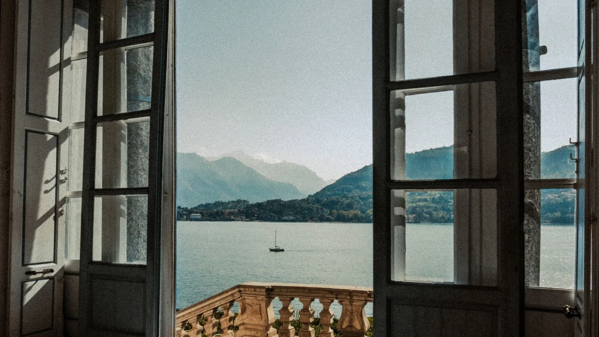 Open window with glass panes framing a view of a lake, mountains in the background, and a sailboat on the water, with a decorative stone balcony in the foreground.