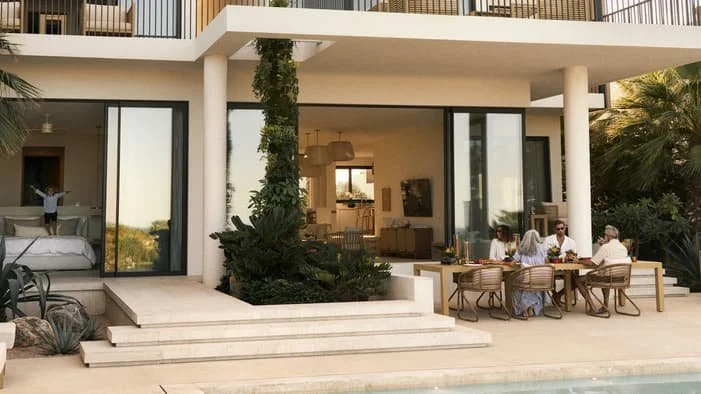 People dining outdoors on a patio in front of a modern house with large glass sliding doors.