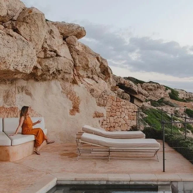 A woman in an orange dress sitting on a built-in bench beside a pool, with a rocky cliff in the background and two lounge chairs nearby.