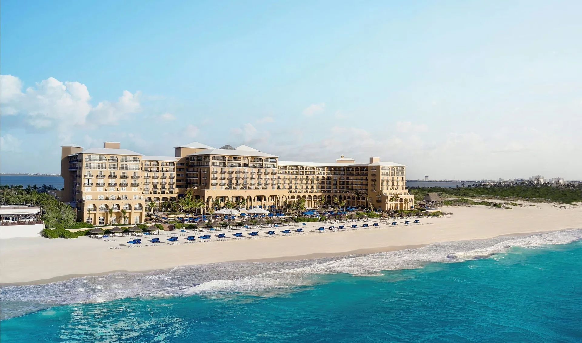A large beachfront hotel with multiple floors and balconies, situated along a sandy beach with turquoise ocean water and blue beach chairs with umbrellas in the foreground, under a partly cloudy sky.