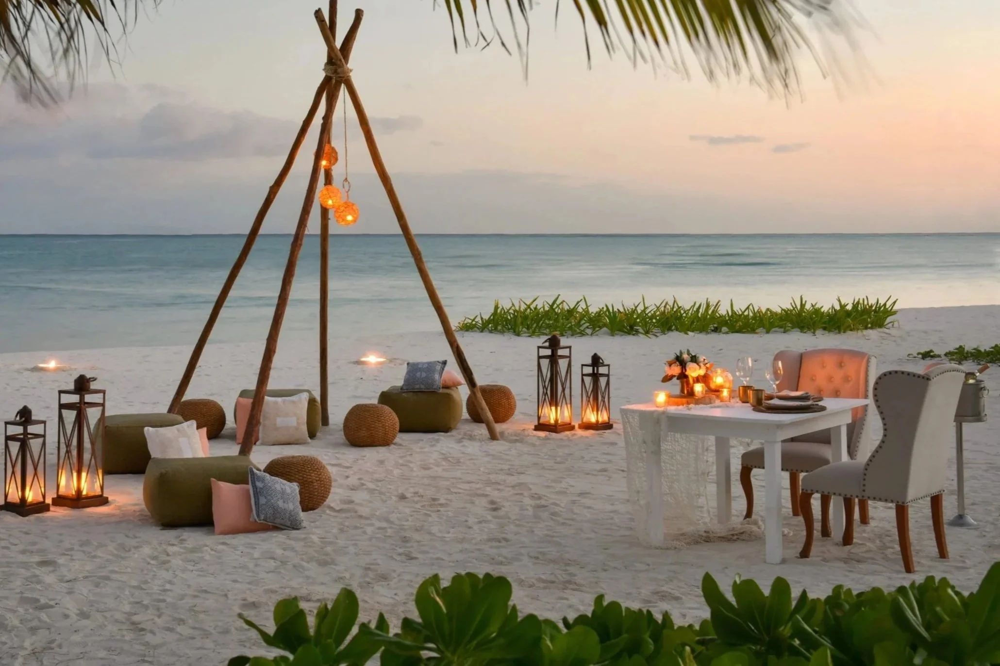 Dinner setup on a beach at sunset with string lights, lanterns, a white wooden table, and chairs, surrounded by sand and water.