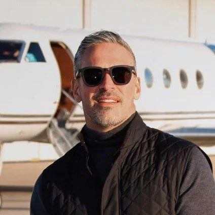 Man with gray hair and beard wearing sunglasses and a black vest, standing in front of a private jet.