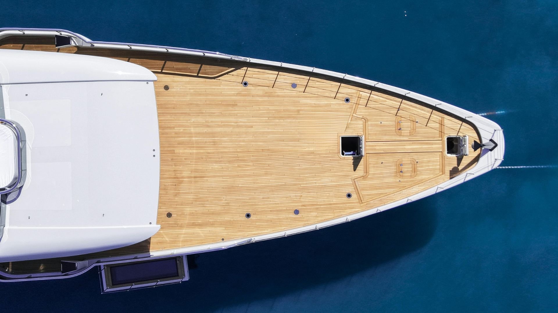 Top-down view of a yacht's wooden deck with two hatches, black fittings, and a white structure on the left, floating on dark blue water.