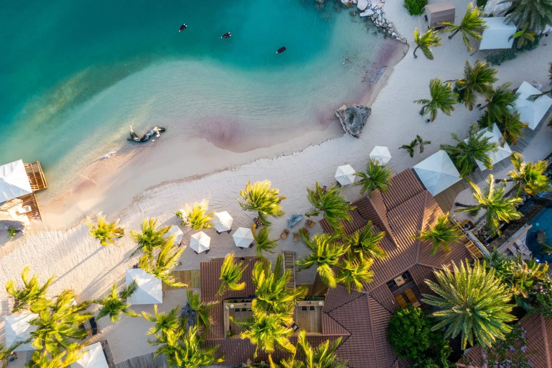 Aerial view of a tropical beach with white sand, clear turquoise water, palm trees, umbrellas, and beachfront houses with red roofs.