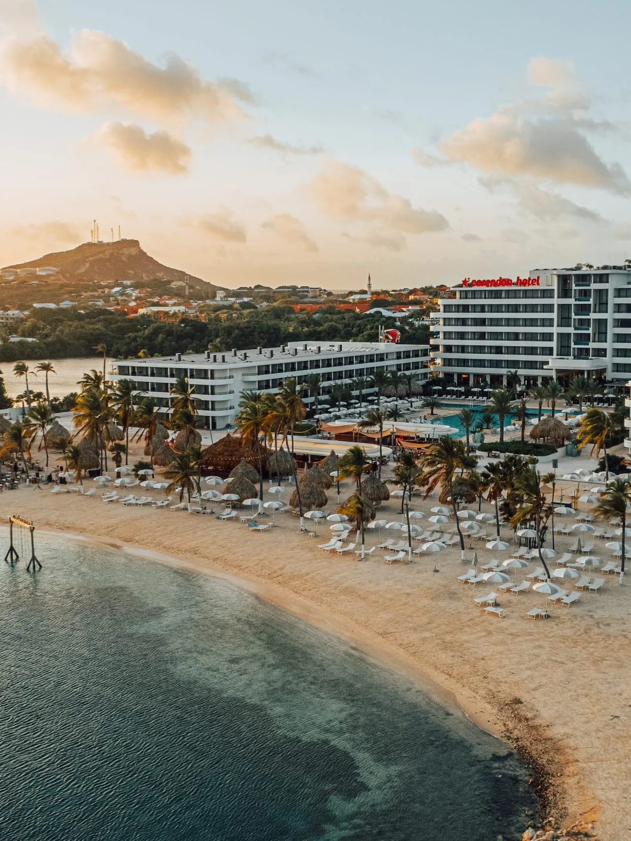 A beachside resort with white lounge chairs and palm trees on sandy shore, a hotel complex with pool, and a hillside with houses and communication towers.