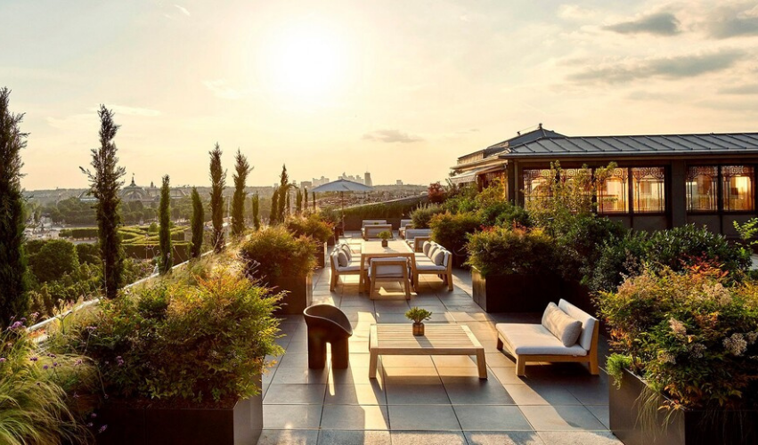 Rooftop terrace with seating area, potted plants, and city skyline in the background during sunset.