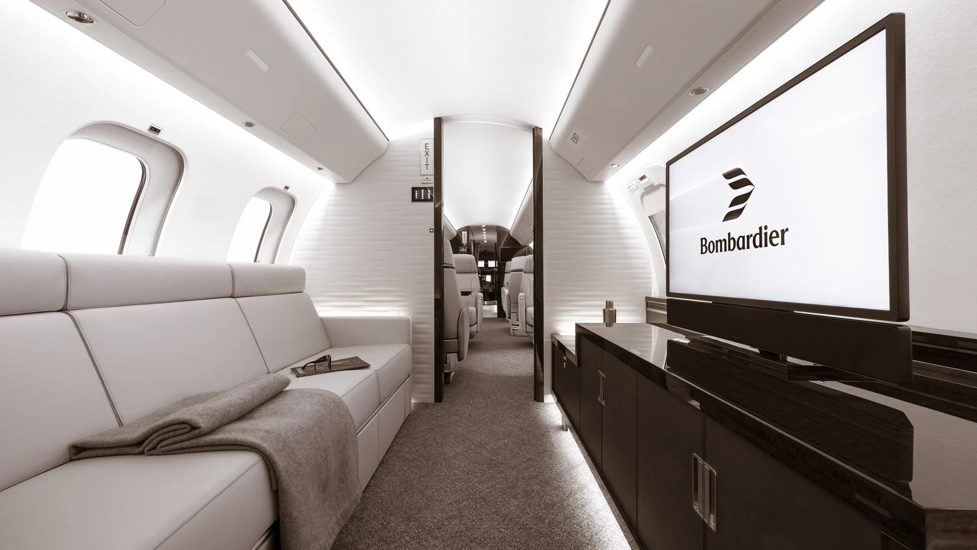 Interior of a private jet with white leather seats, a large screen displaying 'Bombardier', and a hallway leading to additional seating.