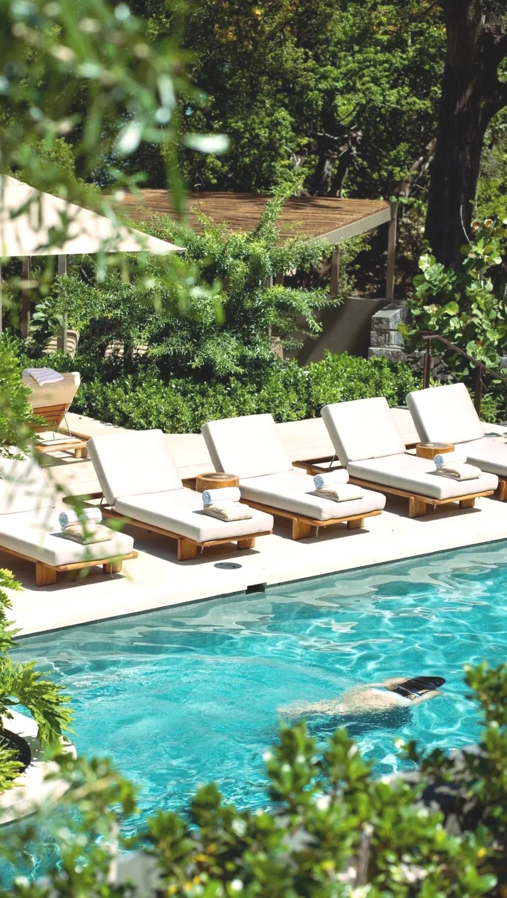 Luxury pool area with four white lounge chairs by the water, surrounded by green bushes and trees, with a person swimming in the pool.