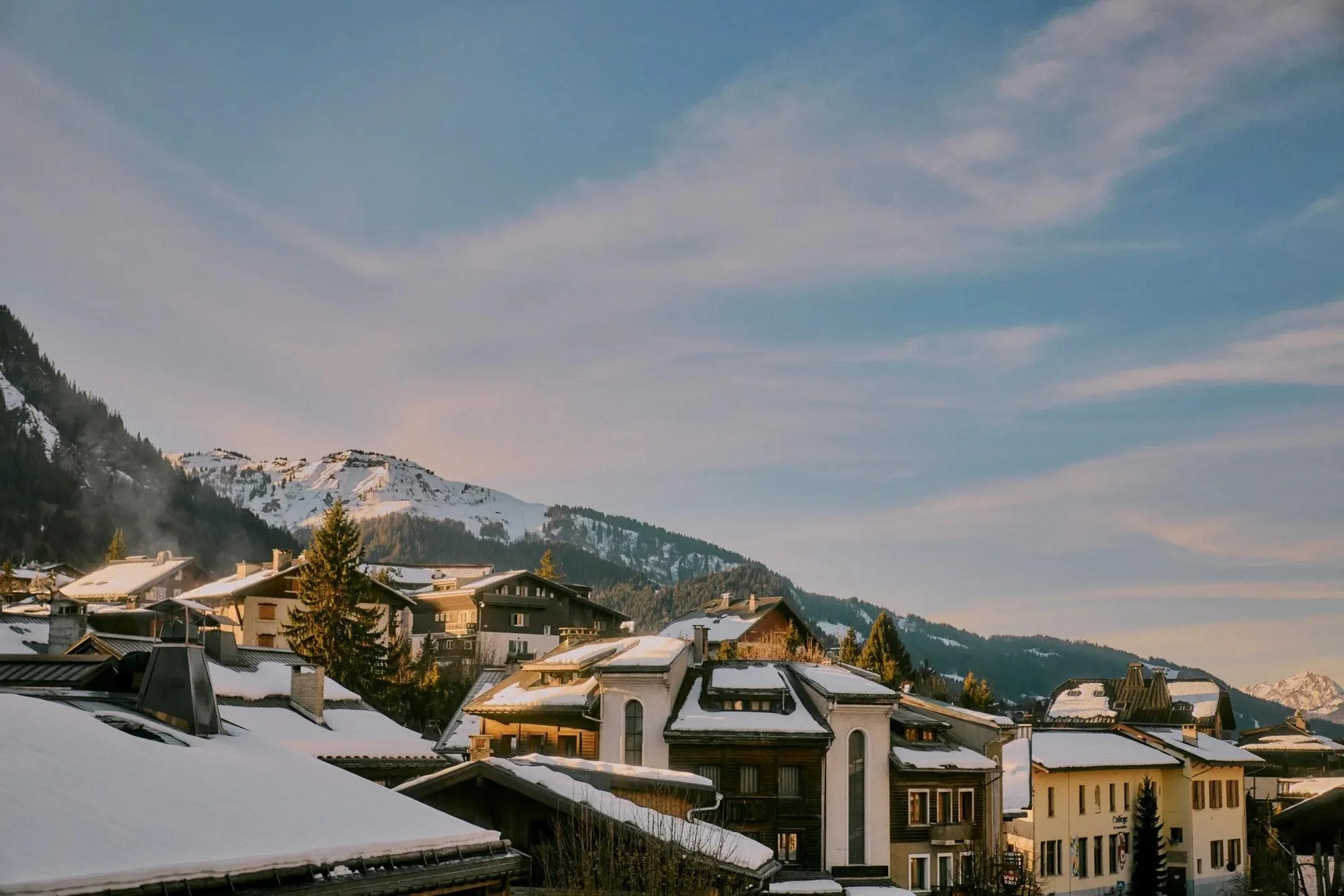 Snow-covered rooftops of houses in a mountain town with a snow-capped mountain in the background under a partly cloudy sky.