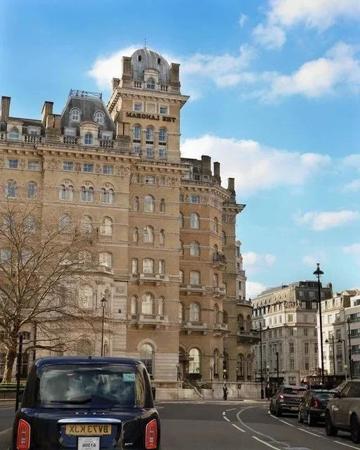 A historic multi-story building with a rounded tower and ornate architecture on a city street, with parked cars and a clear blue sky.