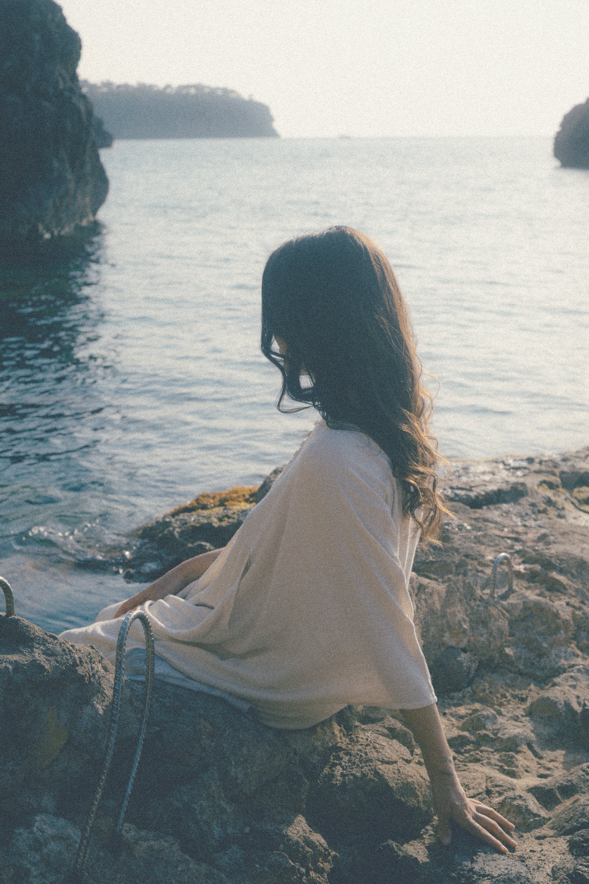 A woman sitting on rocks by the water, facing the sea, during sunset or sunrise.
