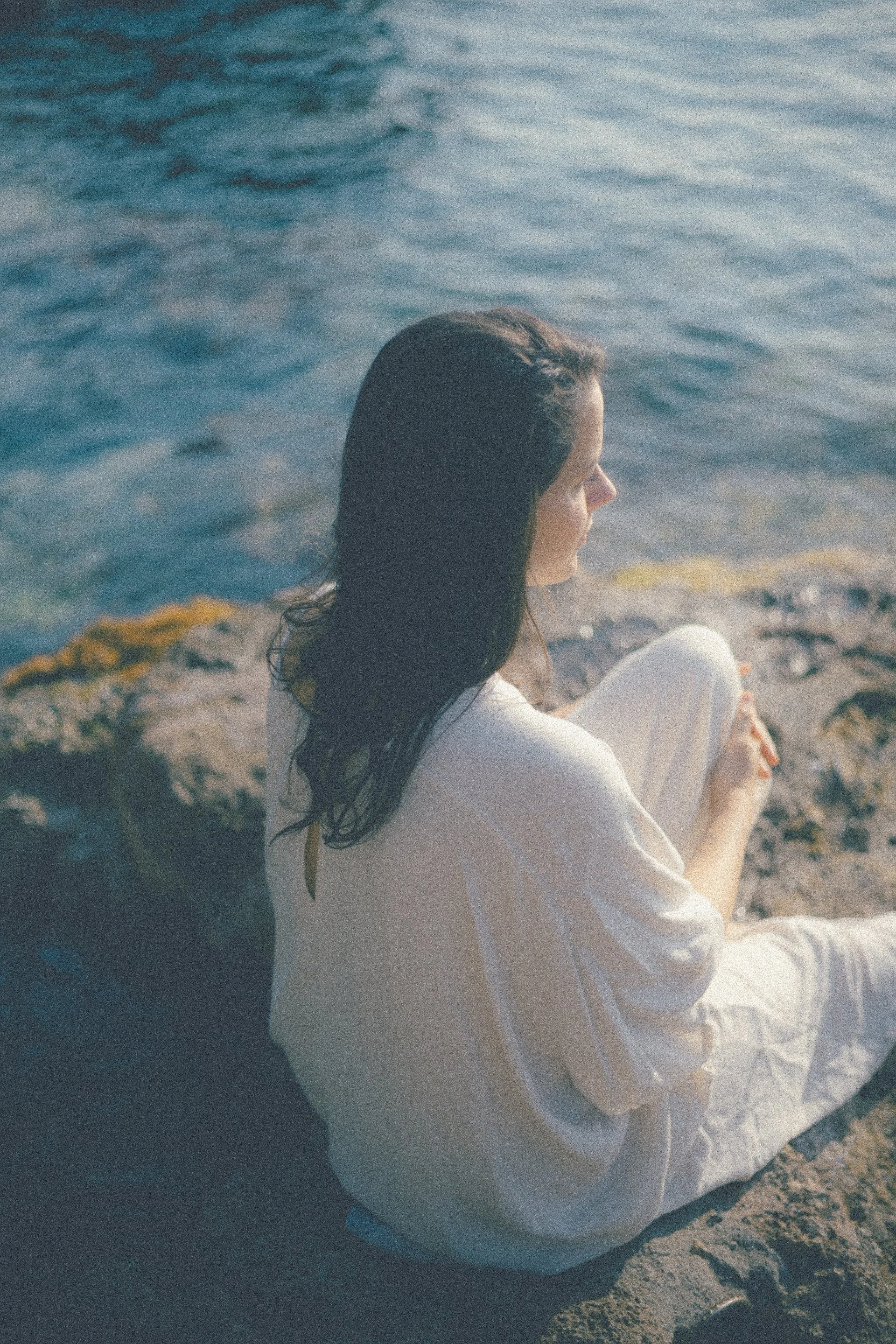 A woman with dark hair sitting on rocks by the water, looking out at the ocean.