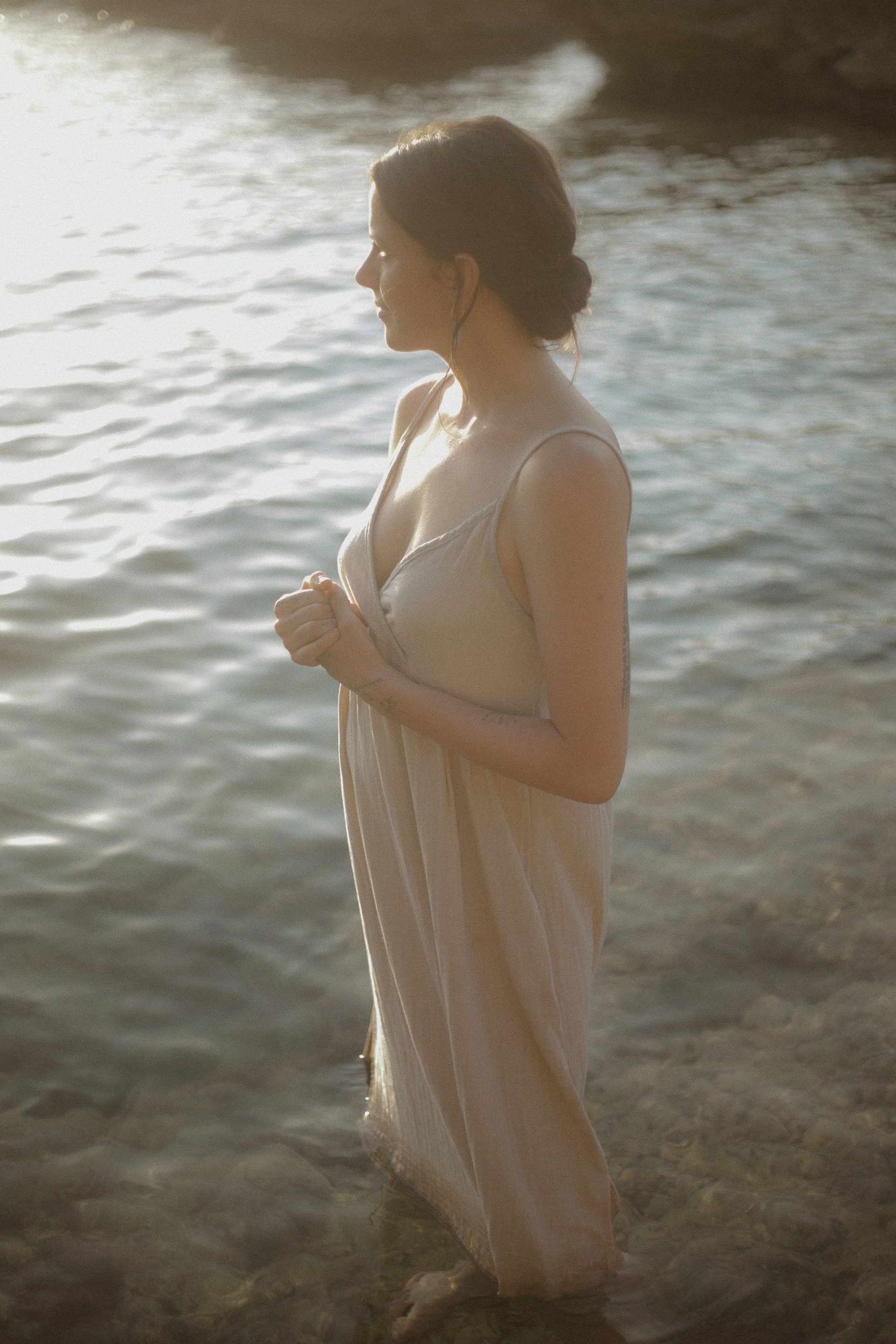 A woman standing in shallow water near a shoreline, wearing a light-colored dress, with her eyes closed and hands clasped together.
