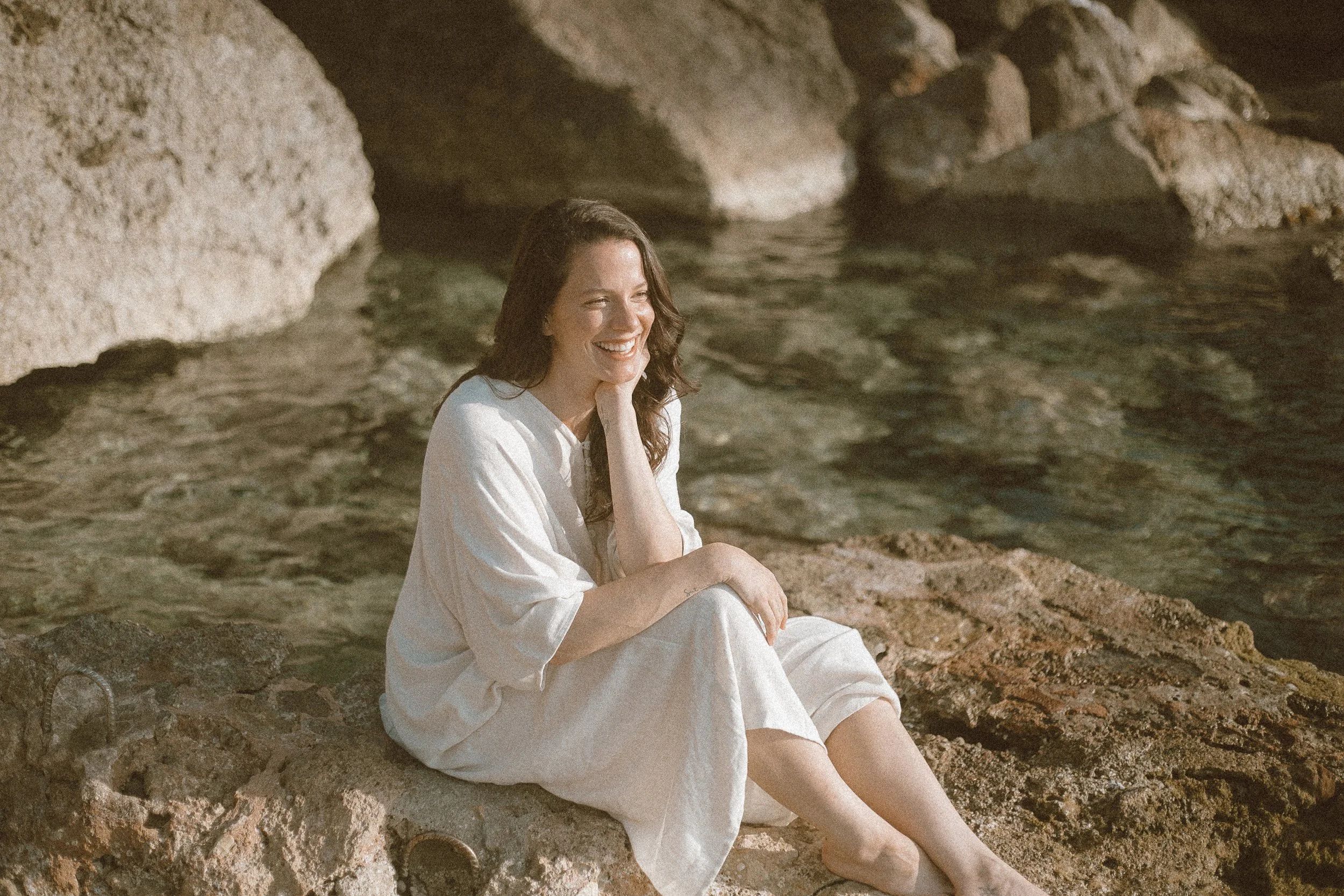 A woman with long dark hair sitting on a large rock by the water, smiling and wearing a white dress.