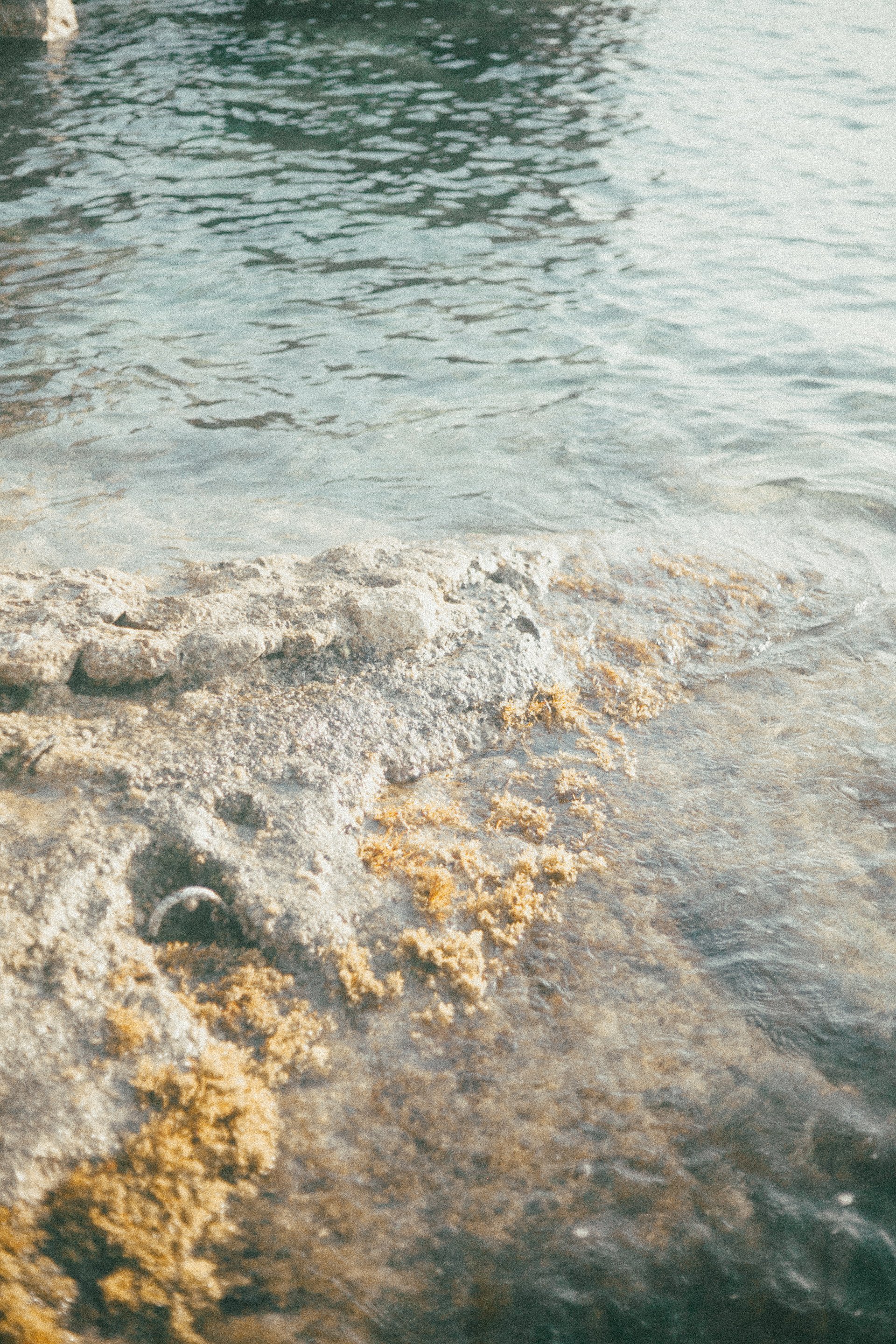 Close-up view of a rocky and sandy shoreline with gentle waves lapping against it, water in the background.
