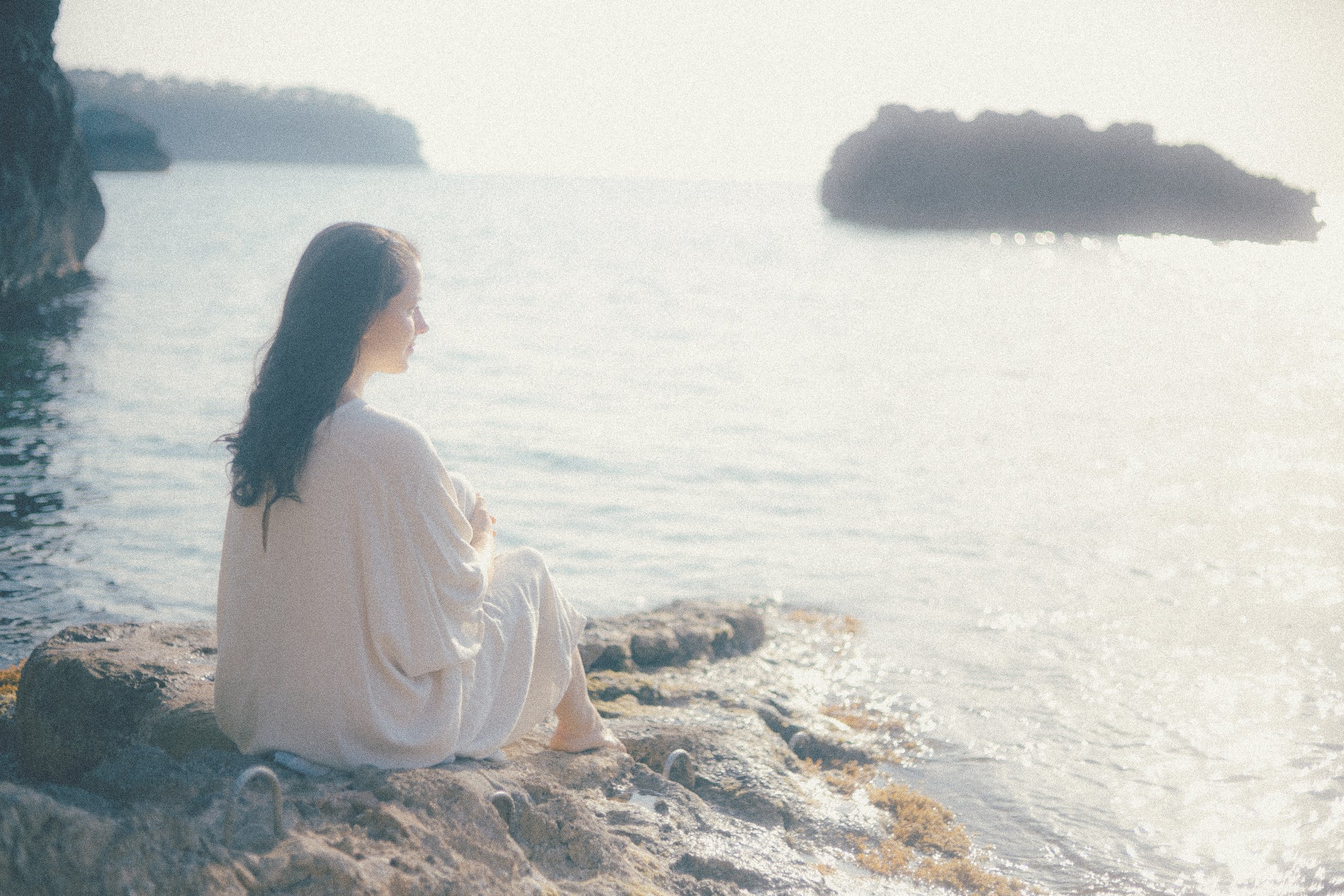 A woman with long dark hair sitting on a rocky shoreline, looking at the water during sunset or sunrise, with distant islands or rocks visible in the background.