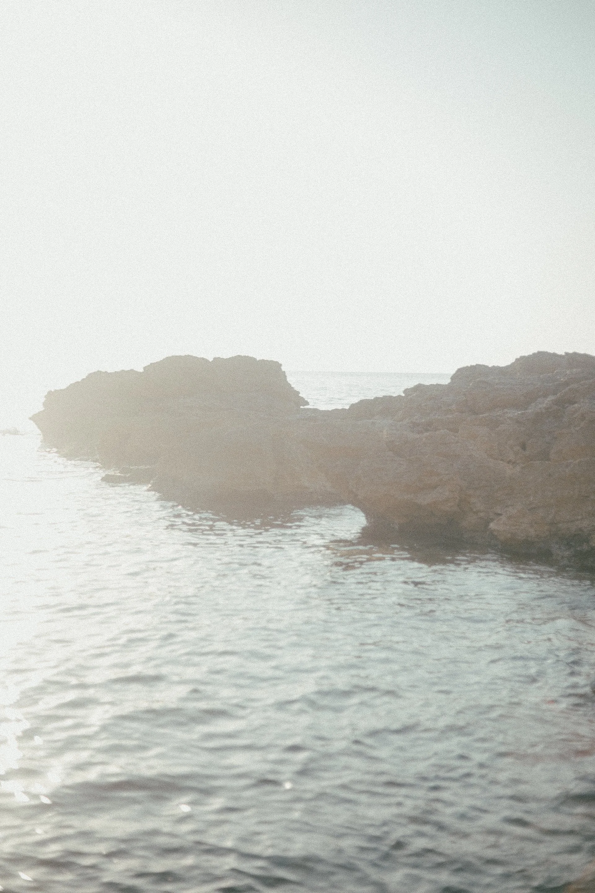 A hazy coastal scene with a rocky shoreline and calm water, possibly at sunset or sunrise.