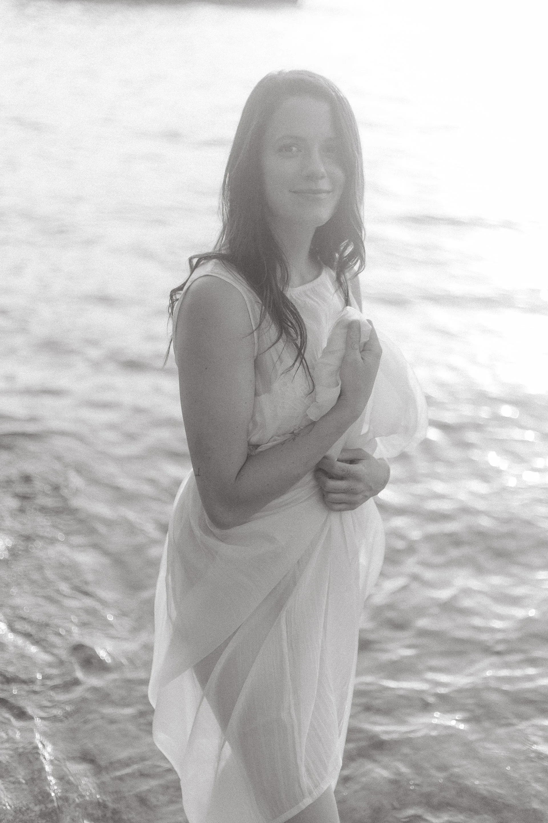 A woman in a flowy dress standing in the ocean, smiling, with wet hair and water around her feet.