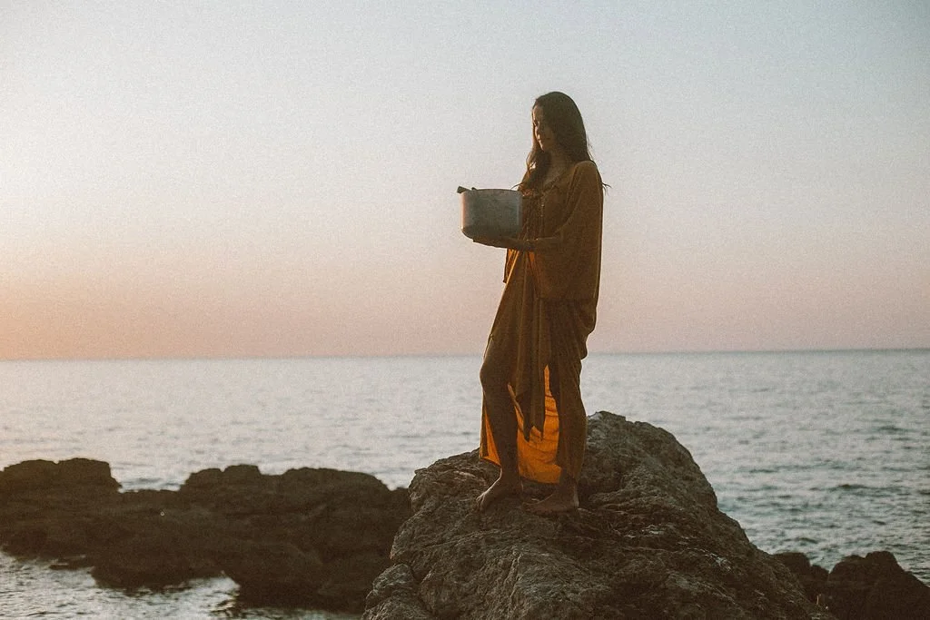 A woman standing barefoot on a large rock by the ocean at sunset, holding a small box.
