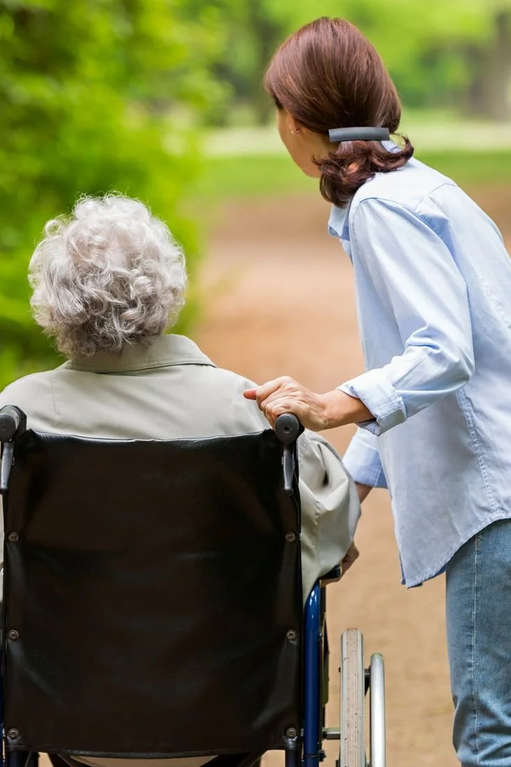 A caregiver pushing an elderly woman in a wheelchair along a nature trail with green trees and a dirt path.