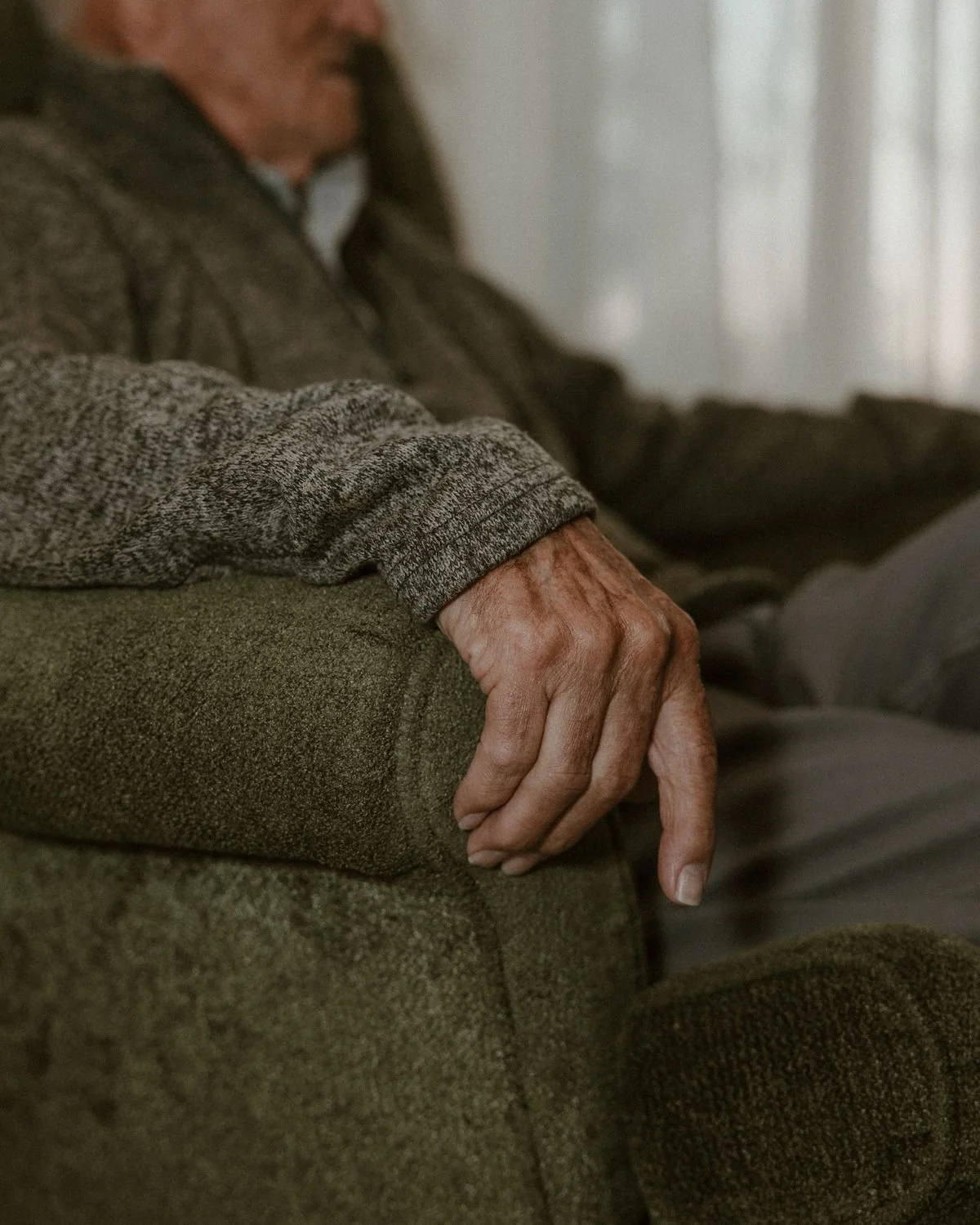 Close-up of an elderly person's hand resting on a cushioned armrest, wearing a gray long-sleeve shirt and sitting on a greenish-brown sofa.