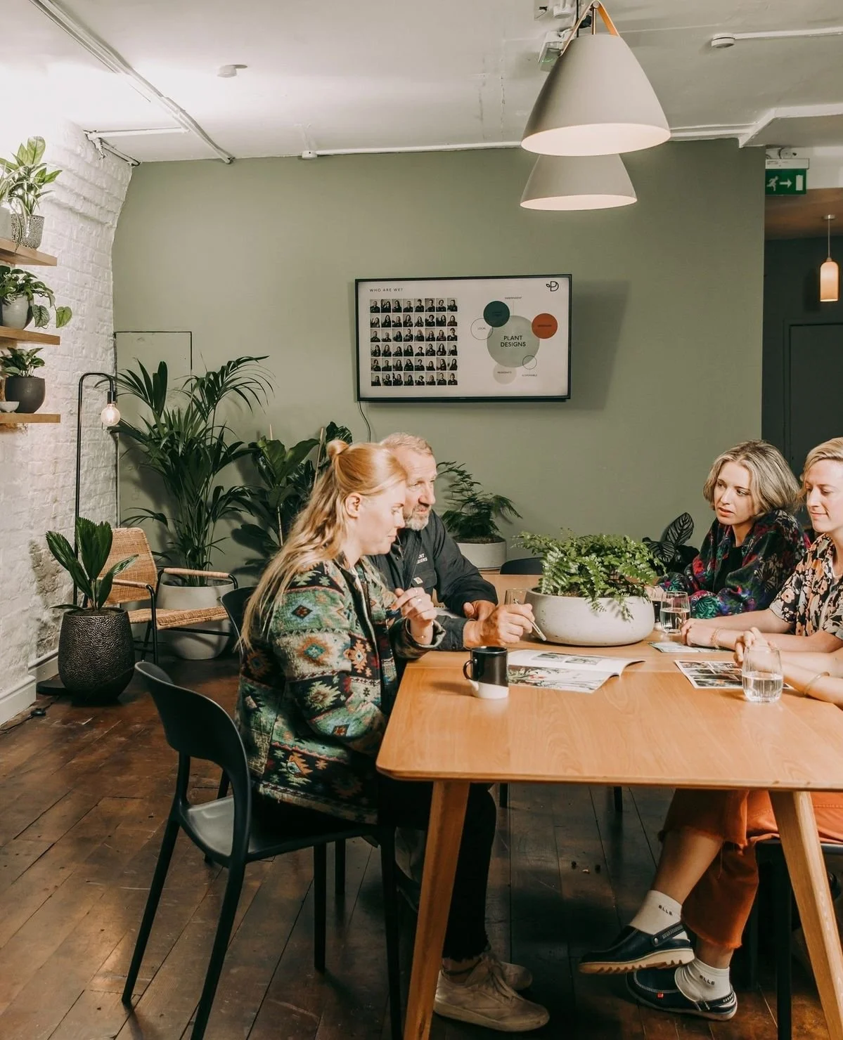 A group of five people sitting at a wooden table in a room with green walls, plants, a wall-mounted monitor, and pendant lighting, engaged in conversation.