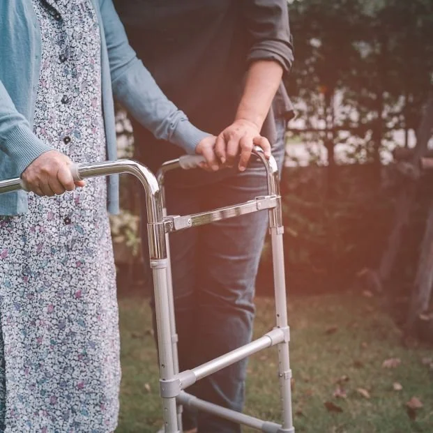 Two people walking outdoors, one with a walker, in a park or garden setting.