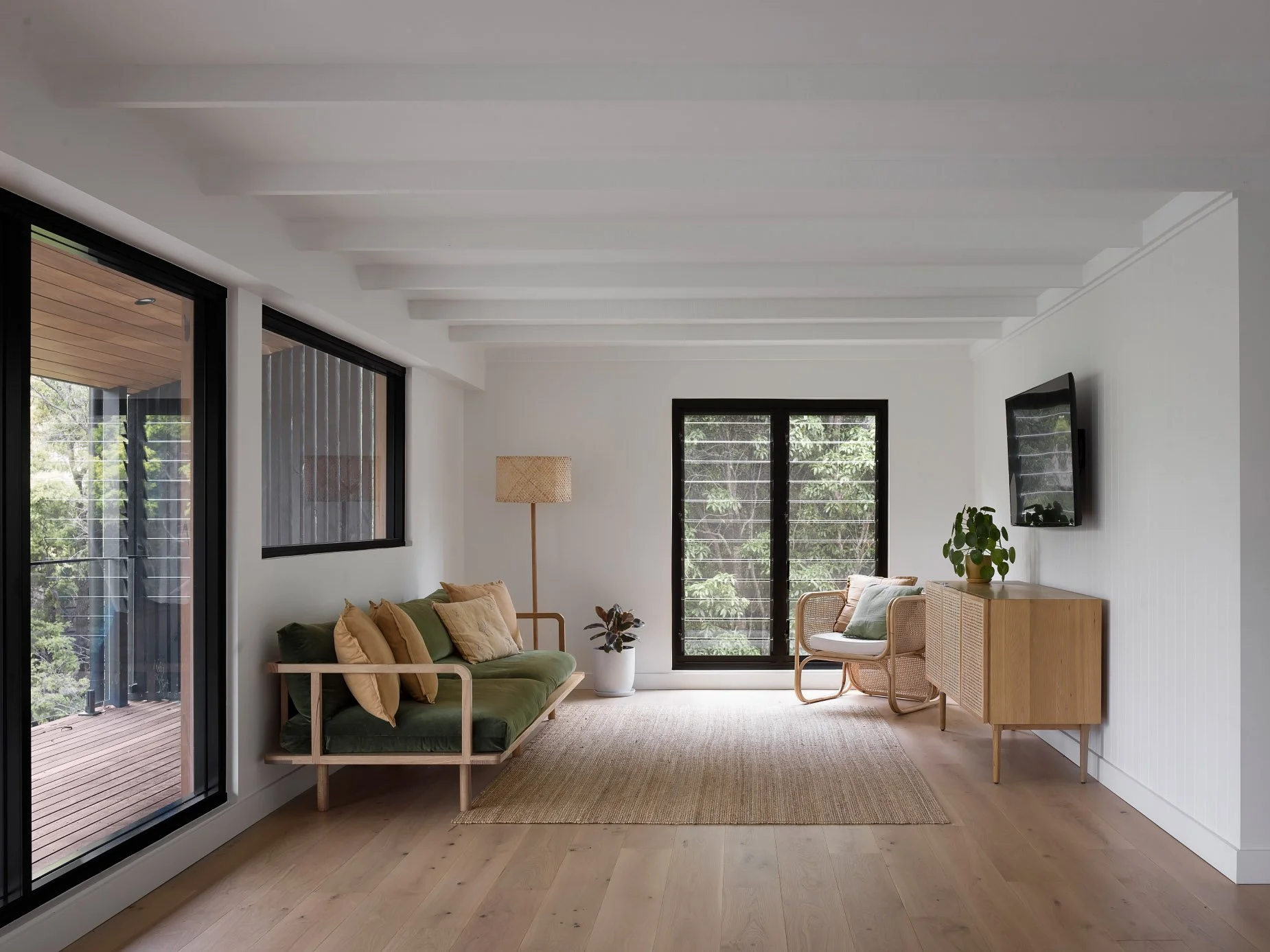 A modern living room with white walls, large glass sliding doors leading to a wooden balcony, a green sofa with beige pillows, a wooden sideboard with a potted plant, a flat-screen TV on the wall, a beige rug, and a rattan chair with cushions. Natura