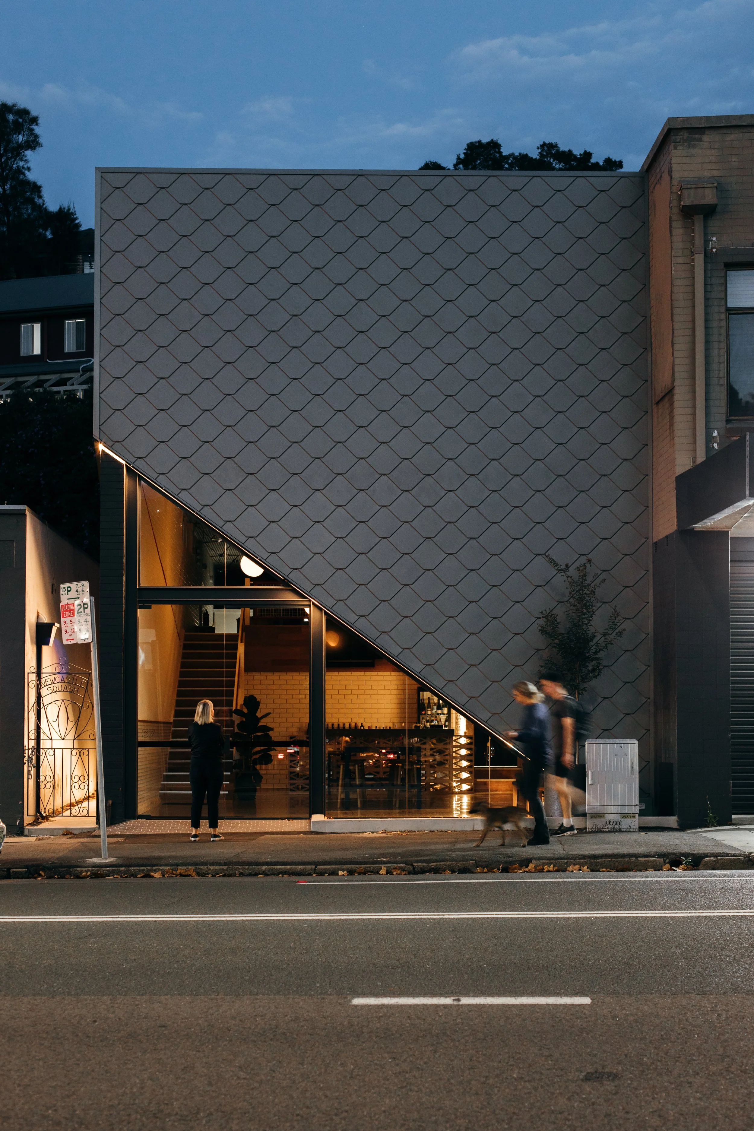 View of a modern building with a zinc cladding exterior, illuminated from within at dusk. People are walking on the sidewalk in front of the building.