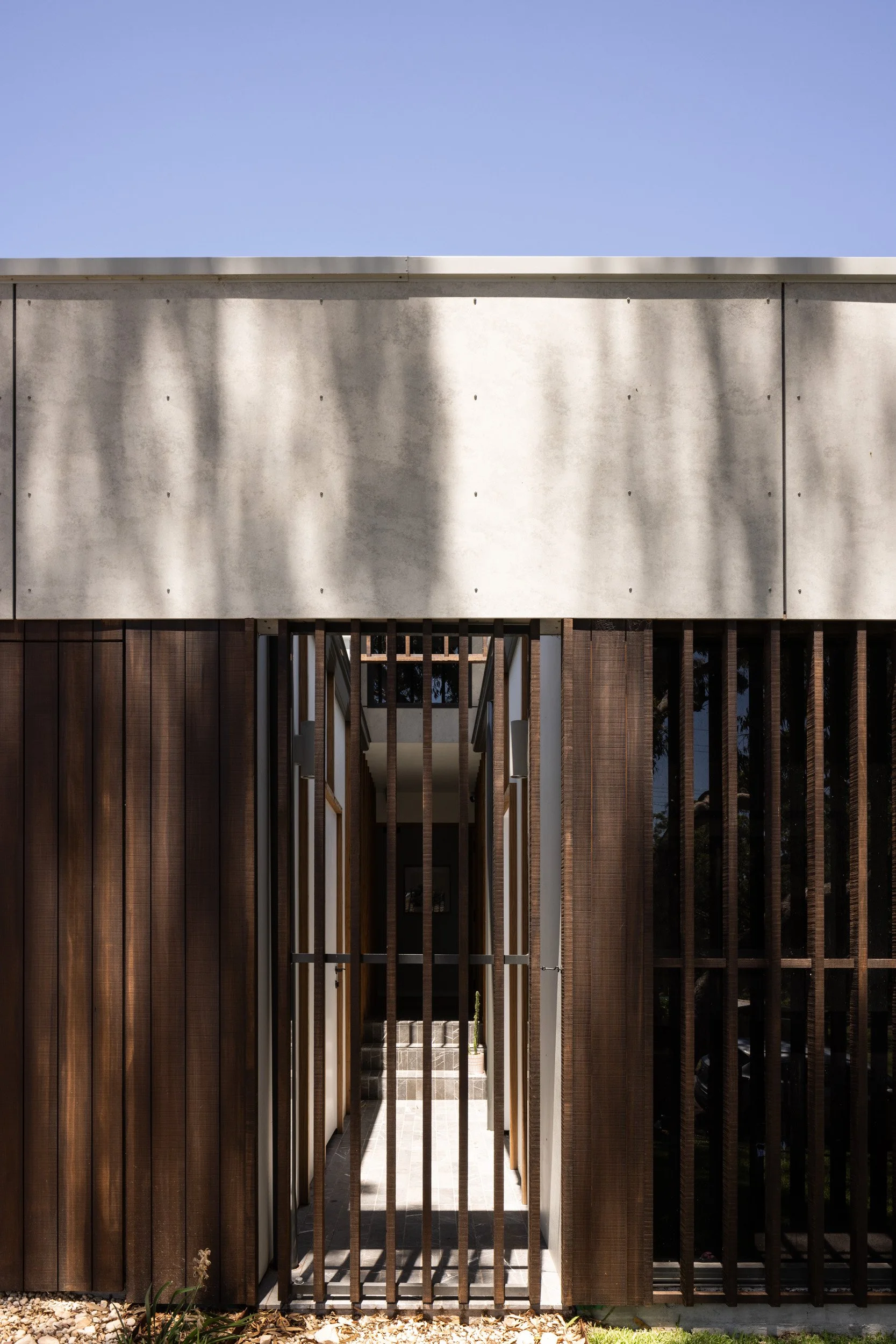 A modern building with vertical wooden slats and a concrete wall, with shadows cast on the wall, and an entrance with a pathway leading inside.