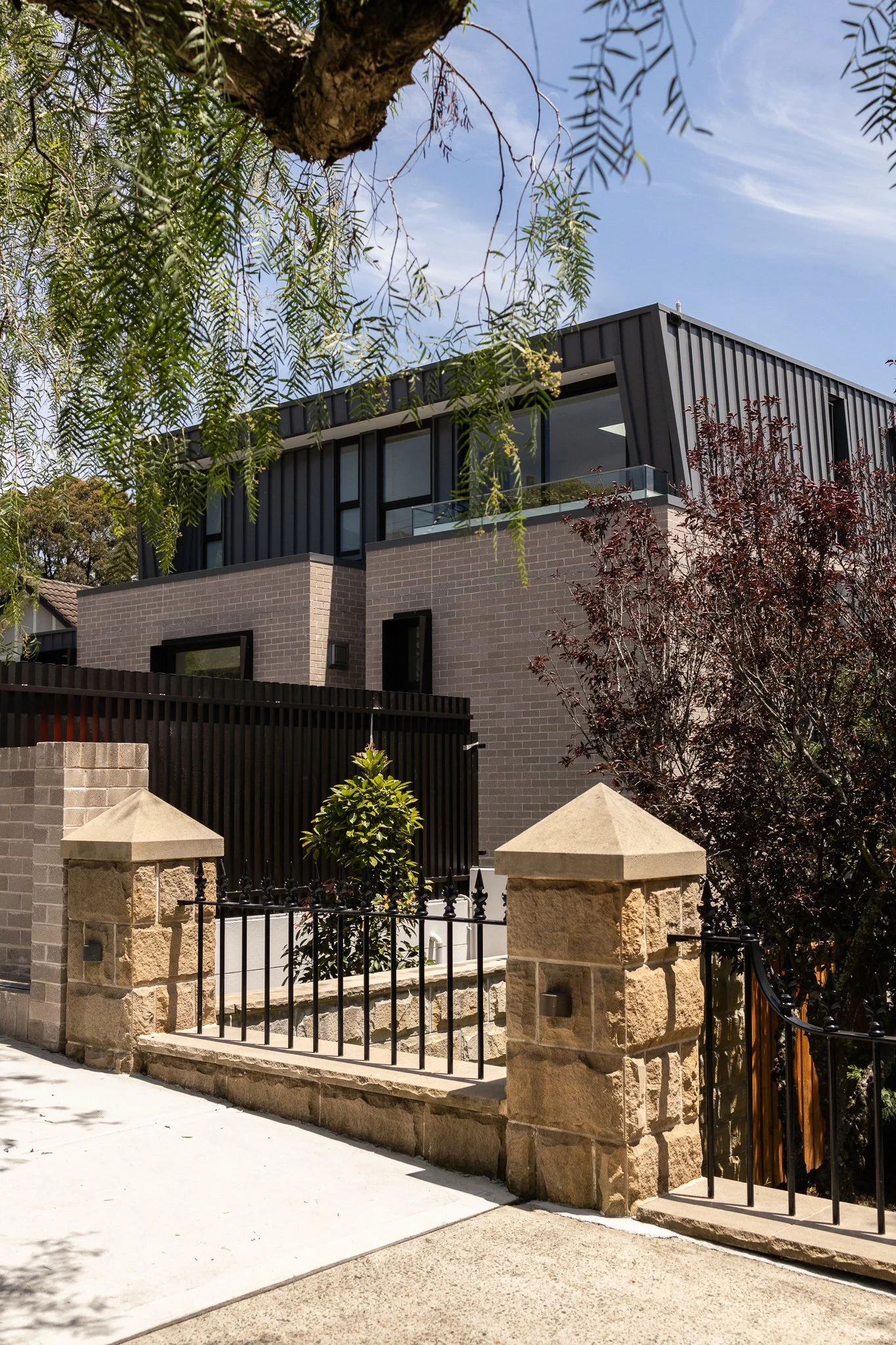 A modern multi-story house with black exterior panels and large windows, surrounded by trees and a stone fence with black iron gate, under a blue sky with some clouds.