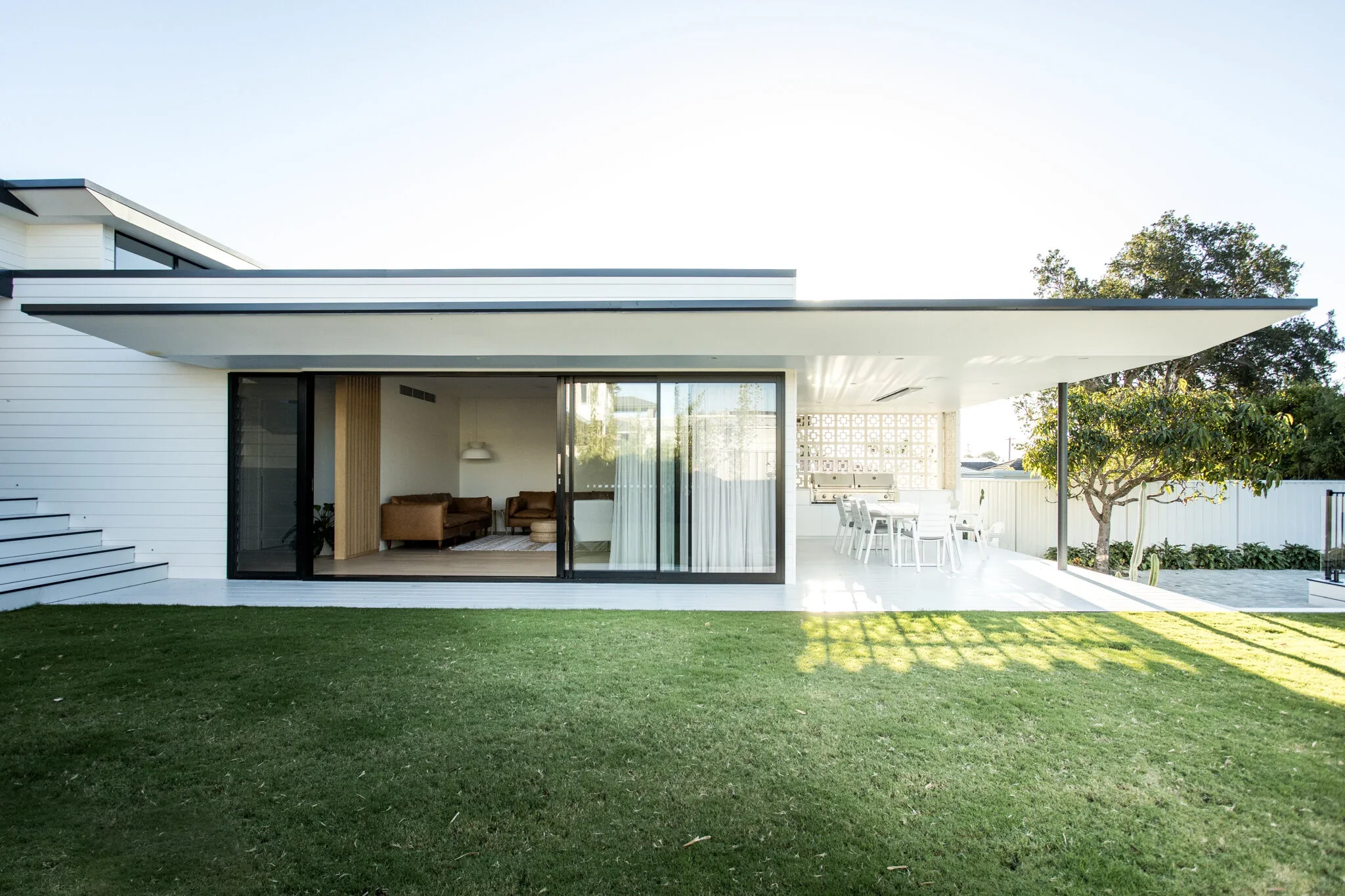 Modern house with large patio, sliding glass doors opening to living room, outdoor dining area with a white table and chairs, green lawn, trees, and a backyard grill area.