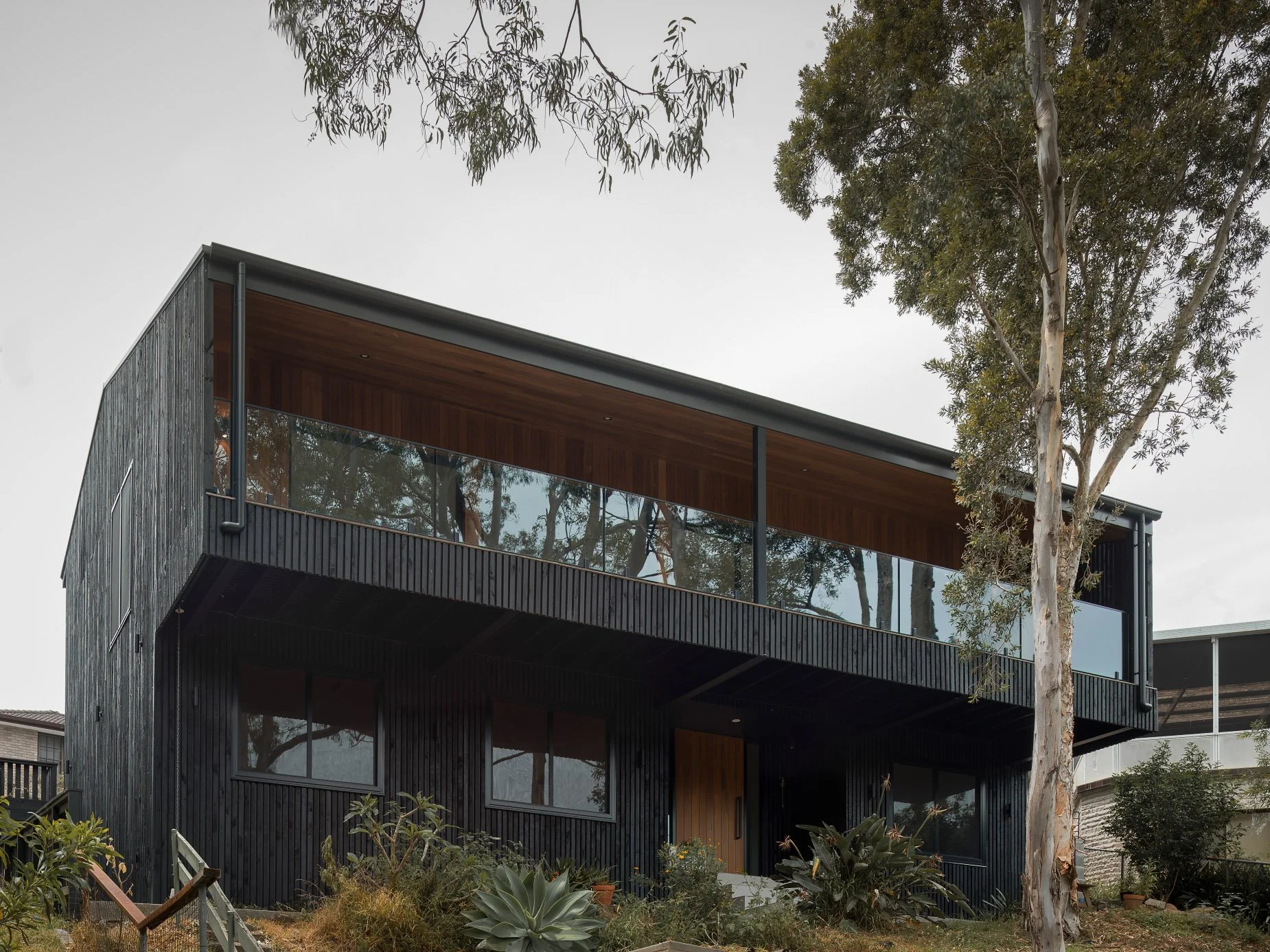 Modern two-story house with dark exterior siding, large windows, and a balcony with glass railings, surrounded by trees and plants.