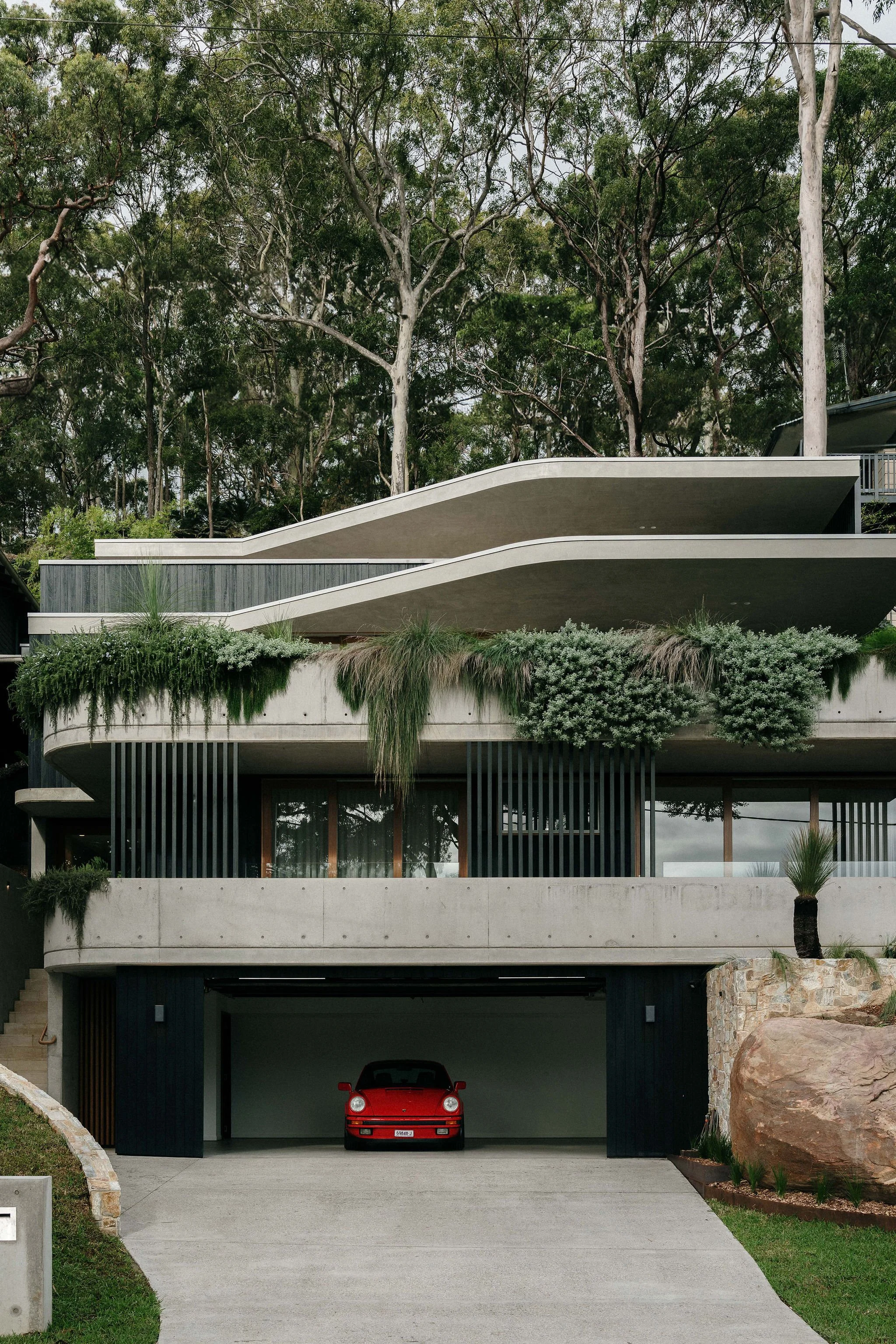 Modern multi-level house with curved off form concrete balconies, greenery, and a red sports car parked in the garage.