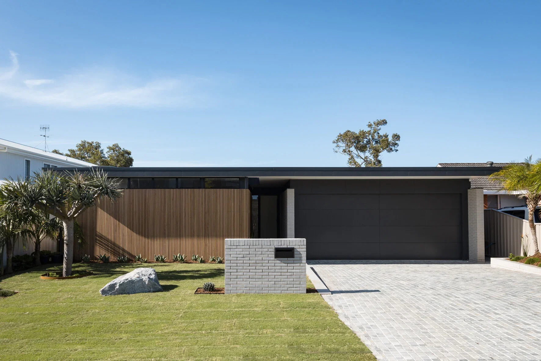 Modern single-story house with a grey garage door, a black roof, a wooden accent wall, and a landscaped front yard with a palm tree, a large rock, and a white brick mailbox.