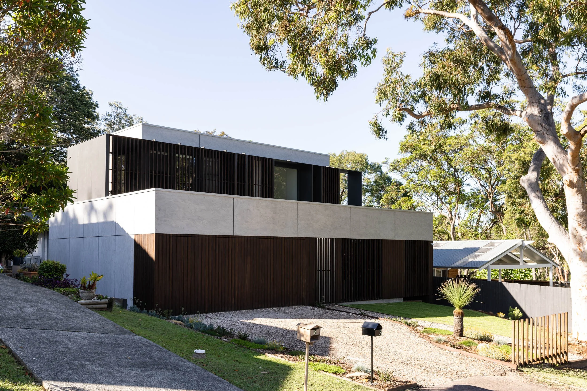 Modern two-story house with white and dark brown exterior, large windows, surrounded by trees and a landscaped yard with a gravel walkway and small plants.