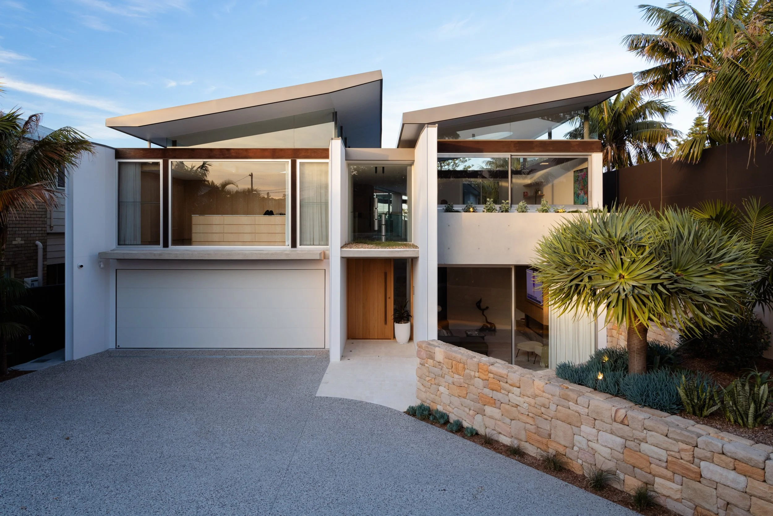 Modern two-story house with large glass windows and a flat roof, surrounded by lush green plants and a brick wall, under a clear blue sky.