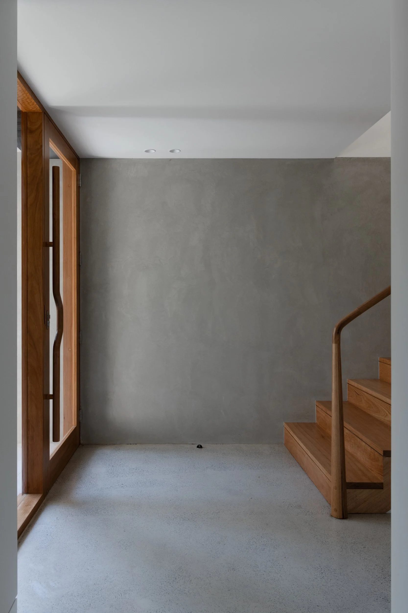 A view of a modern indoor entryway with wooden stairs, a gray wall, a wooden front door with glass panels, and a staircase railing.