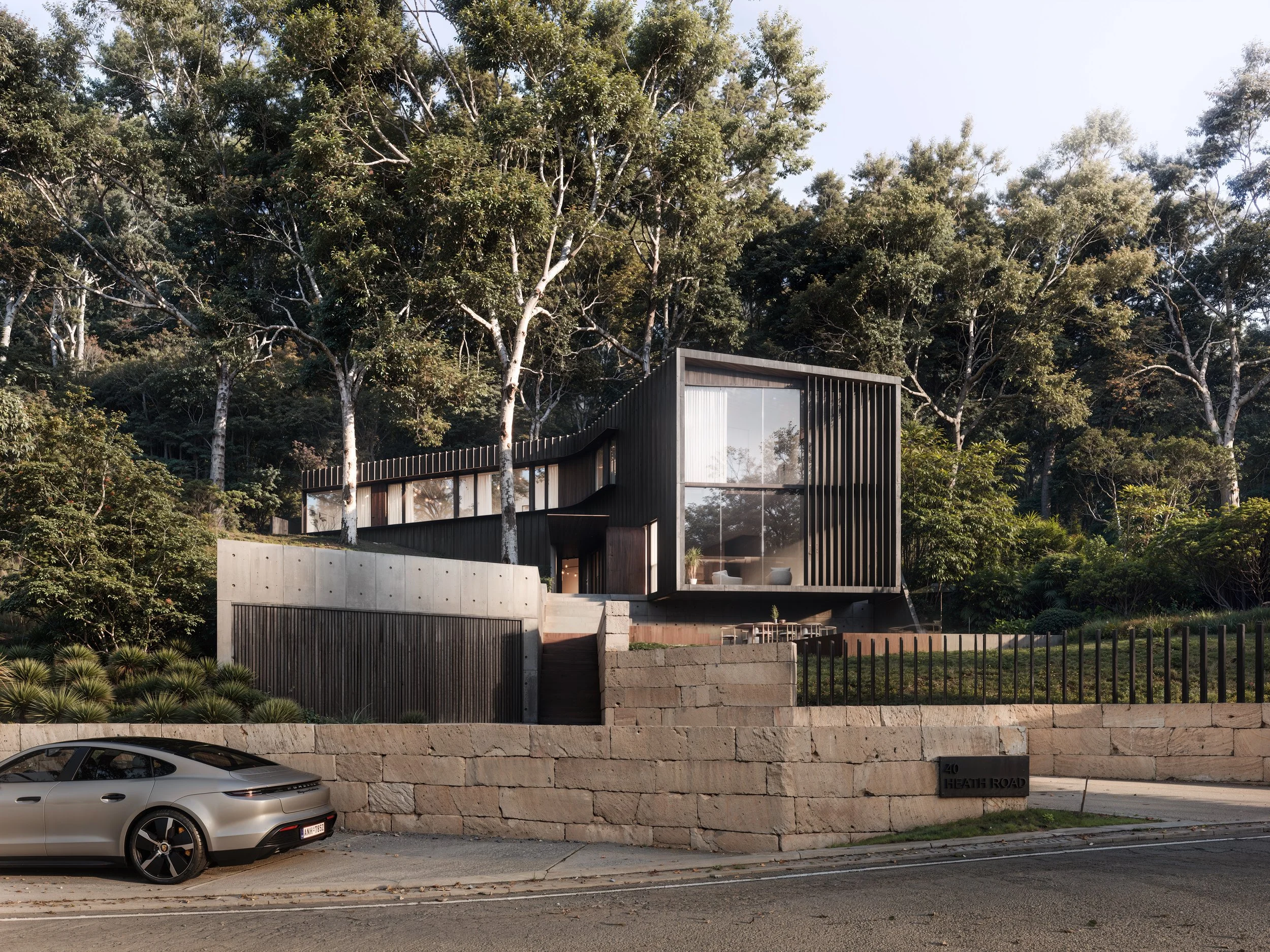 Modern house with large glass windows, black framing, and surrounded by tall trees, located on Heath Road with a stone wall and parked white car in front.