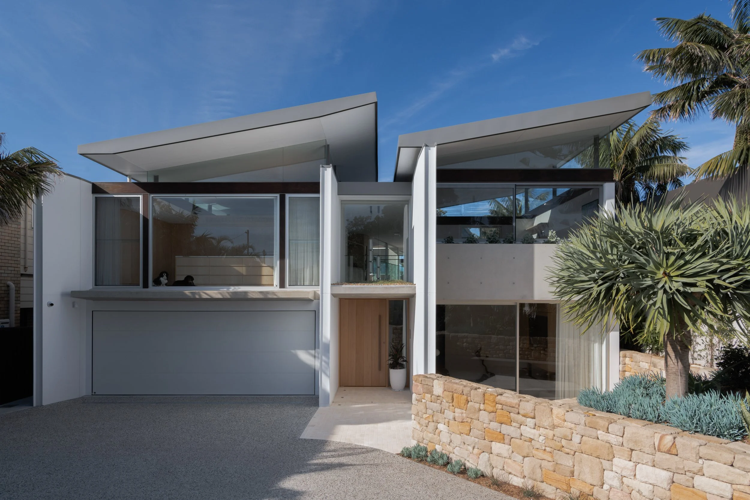 Modern two-story house with large glass windows, a stone wall, and surrounding tropical plants under a blue sky.